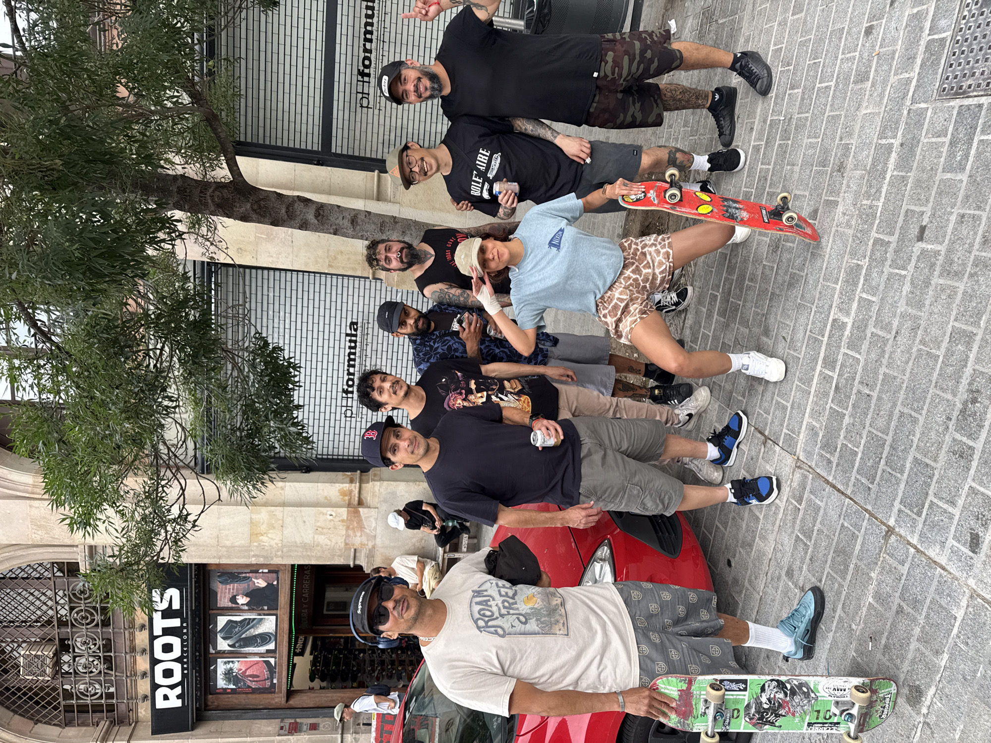 A group of nine men are posing for a photo on a sidewalk in Barcelona, Spain. They are standing in front of a red car parked next to a storefront labeled "ROOTS BARCELONA", which displays images of footwear in its window. A tree is positioned to the left of the group, partially obscuring the building facade and a "pHformula" sign above. Several of the men are holding skateboards. The street is paved with rectangular pavers. The group is in casual attire and engaging with the camera.