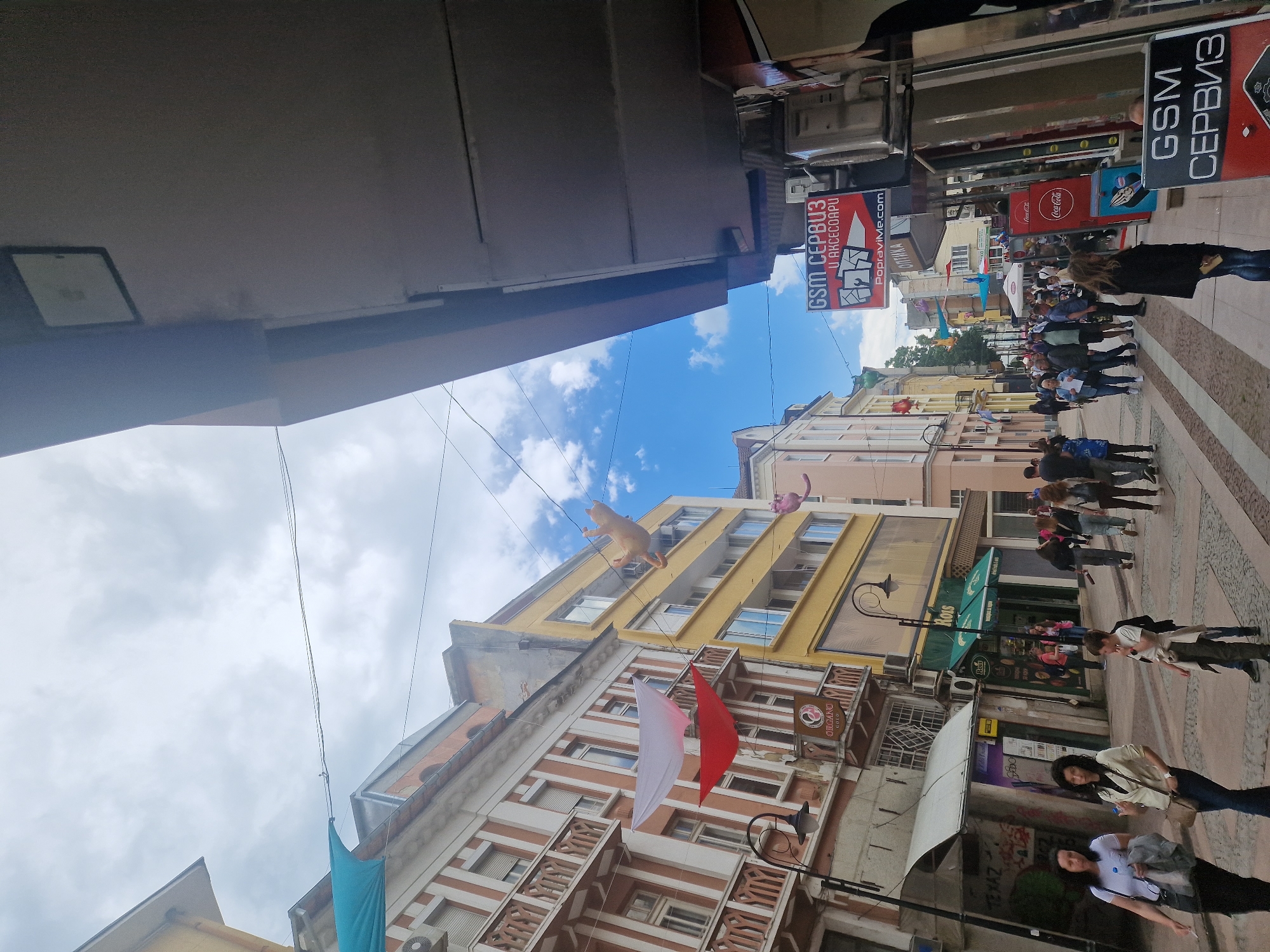 The image depicts a bustling pedestrian street in an unknown city. The street is lined with buildings of varying architectural styles and heights. Several people are walking along the sidewalk, some appearing to be engaged in conversation. Various signs and advertisements are visible on the buildings. Notably, there are two signs for "GSM Сервиз" (GSM Service) in Cyrillic, one with the additional text "U AKCECOAPU" and another with "PopraviMe.com." A Coca-Cola advertisement is also seen. Above the street, inflatable cat-like figures are suspended, and colored fabric banners are hung between buildings. Streetlights are mounted on the sides of buildings.