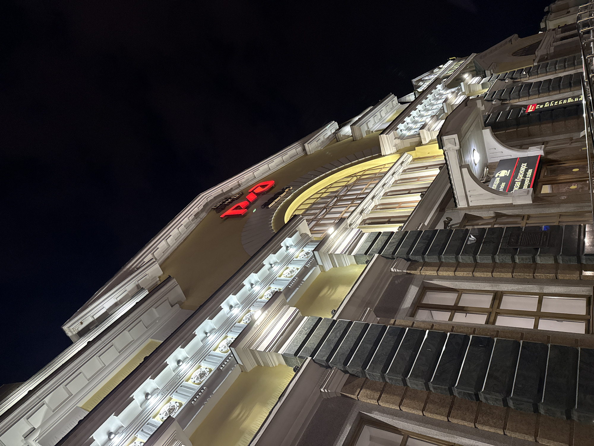 The image shows an upward-angled view of a building at night. The building is adorned with decorative architecture, including illuminated molding and a large, arched window structure. A red sign with the word "RIO" is visible on the facade. A sign with Russian writing indicates the building is near the Krasnoyarsk Railway Station. The building appears to be located in Krasnoyarsk, Russia. The sky is dark and mostly empty. The perspective is from street level looking up at the building.