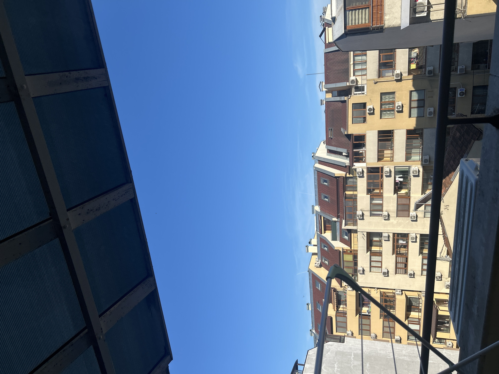 The image shows a view of residential buildings from a balcony, with a clear blue sky above. The buildings are of various heights and architectural styles, with red tiled roofs and yellow facades. Many of the buildings have multiple air conditioning units mounted on their exteriors. There are also windows and balconies visible on the buildings, some with laundry hanging out to dry. In the foreground, a metal railing and a portion of a covered structure are visible, suggesting the vantage point is from a balcony or similar elevated outdoor space. The scene appears to be set in the city of Varna, Bulgaria, judging by the characteristic architecture and climate depicted. There are no people or animals visible in the image.