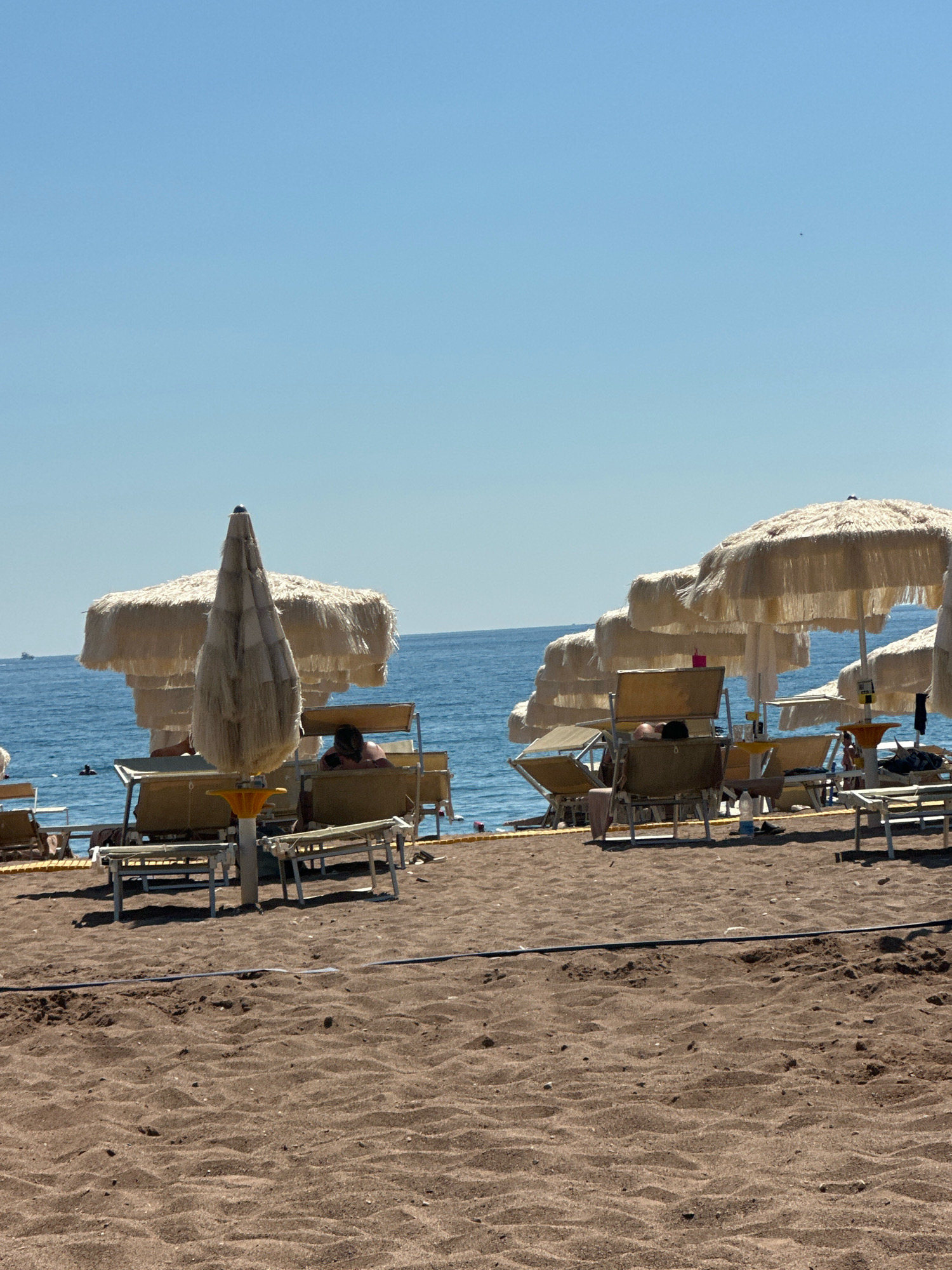 The image depicts a beach scene in Boreti, Montenegro, under a clear blue sky. The foreground is dominated by dark brown sand, with subtle undulations and scattered small pebbles. A dark, thin cable runs horizontally across the sand in the mid-ground.
In the mid-ground, a row of beach chairs and umbrellas is arranged. Several beige beach umbrellas with fringed edges provide shade. Some of these umbrellas are partially folded or in use. Beneath them, lounge chairs are visible, some occupied by people. On the left, a large, textured umbrella is prominent, its woven material and layered fringes clearly visible. Two individuals are seated on lounge chairs behind this umbrella, one appearing to be resting.
Further back, the blue sea stretches to the horizon, meeting the sky in a distinct line. A faint, distant ship is visible on the left side of the sea. The water appears calm. The beach appears to be part of a resort or public beach facility, indicated by the consistent arrangement of seating and umbrellas. The overall atmosphere is one of leisure and relaxation.
