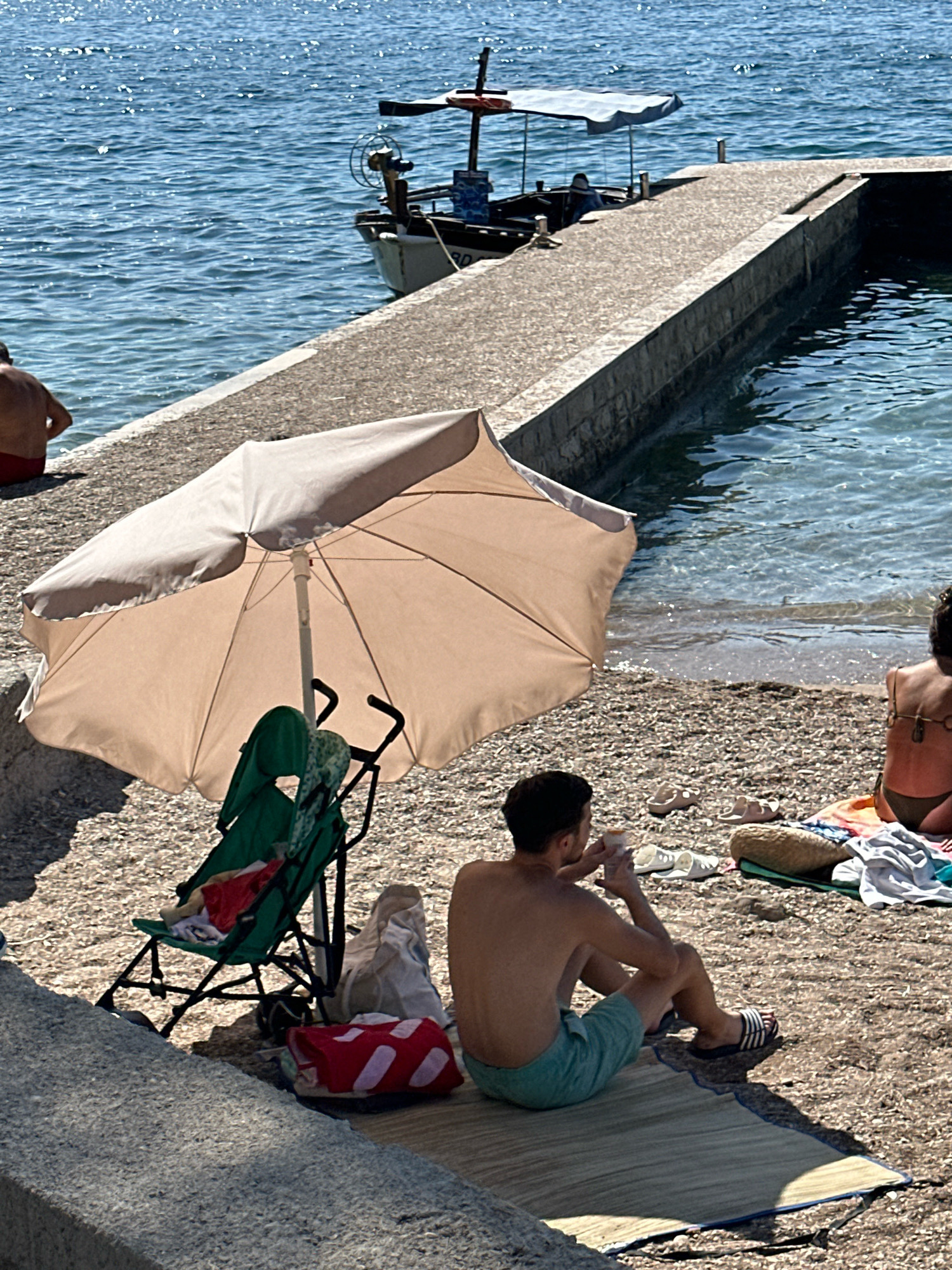 The image captures a sunny day at a beach in Budva, Montenegro. A man is seated on a mat on the pebble beach, facing away from the camera, holding a drink. He is wearing blue shorts and striped sandals. Beside him, a green baby stroller is partially visible under a large, light-colored beach umbrella. A red and white bag rests on the ground near the stroller. In the background, a concrete pier extends into the clear blue water, where a small boat is docked. Another person, partially visible, is seated further down the beach, facing the water. The sea sparkles under the bright sun. The overall scene depicts a relaxed beach atmosphere.