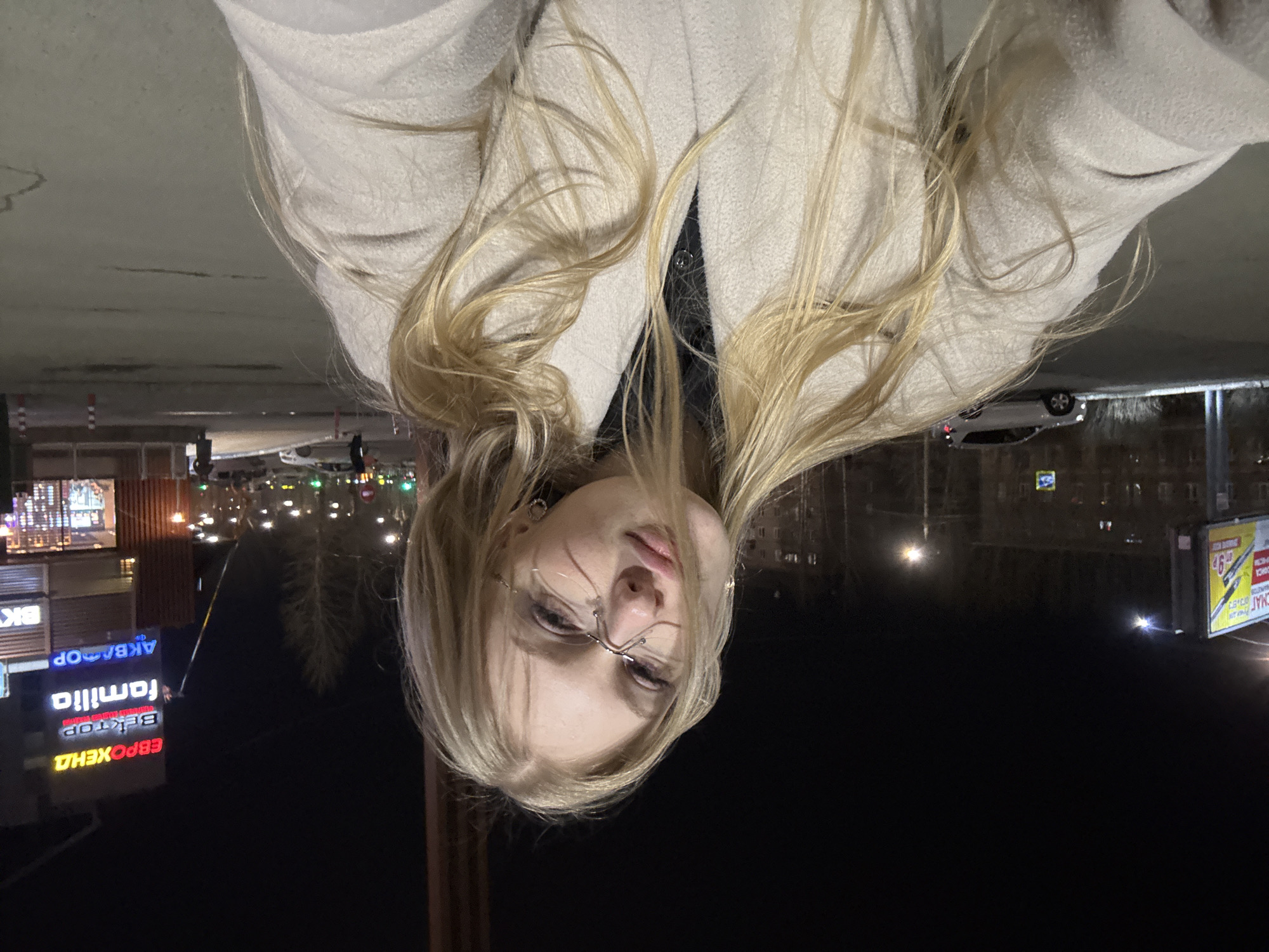 A young woman with blonde hair is shown in an upside-down outdoor selfie. She is wearing glasses and a light-colored coat. The background is dark, indicating nighttime, with illuminated signs in Russian visible. Signage includes "AKBAOOP," "familia," "BEKTOP," and "EBPOXEHA." The location appears to be a city street in Perm, Russia, based on the signage and architectural style. Street lights and buildings are visible in the background, along with a car.