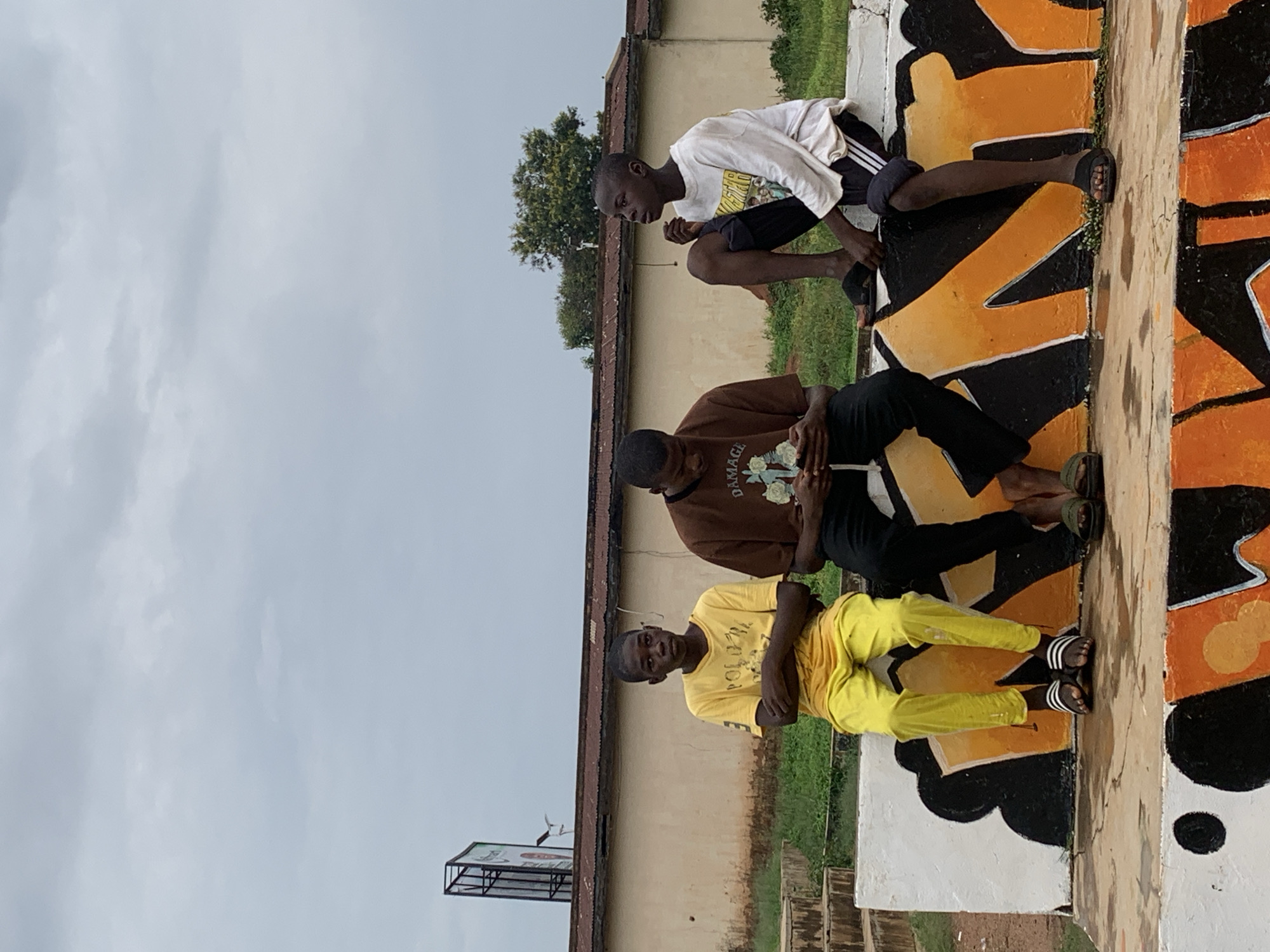 Three young Black males are sitting on a concrete structure outdoors, likely in Jos, Nigeria, given the context. The individual on the left is wearing a bright yellow t-shirt and yellow pants, with his arms crossed and looking directly at the camera. The middle individual is wearing a brown t-shirt and black pants, looking down at his phone. The individual on the right is wearing a white long-sleeved shirt and black pants, looking off to the side. They are all wearing casual footwear. The concrete structure they are sitting on is decorated with graffiti in orange and black. Behind them, there is a building with a corrugated iron roof, some green grass, and trees. A billboard structure is visible in the distance to the left. The sky is overcast.