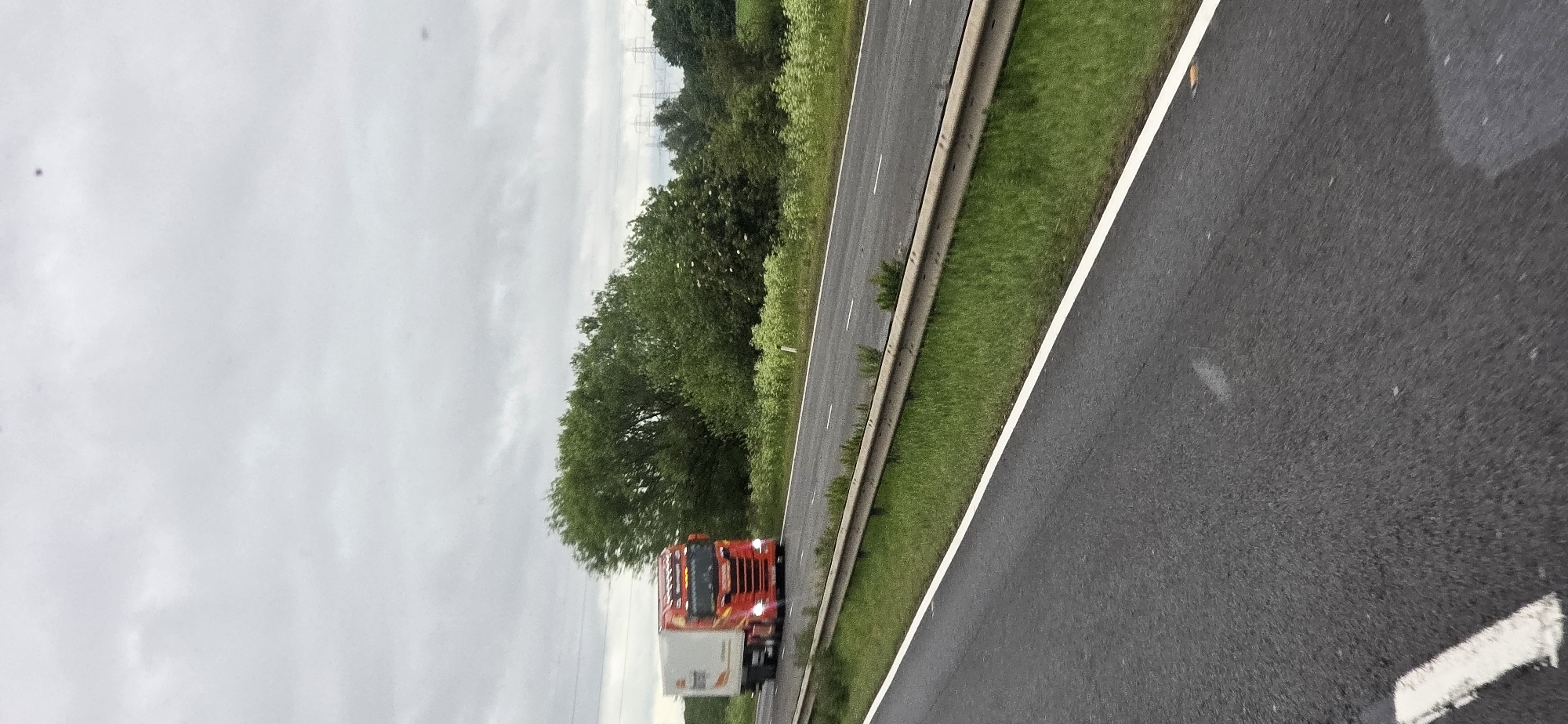 The image shows a red and white articulated lorry in motion on a multi-lane highway. The lorry is positioned on the left lane, with its headlights illuminated. The background consists of lush green trees and vegetation, suggesting a rural or semi-rural setting. In the distance, there are tall electricity pylons visible against a cloudy sky. The highway itself has white lane markings and a metal guardrail separating the road from the grassy verge. The perspective suggests the photo was taken from another moving vehicle on the same highway, looking towards the lorry. The overall impression is one of a typical daytime journey on a major road.