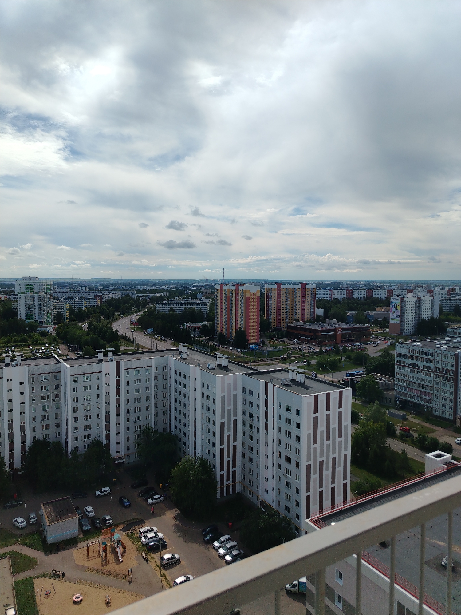 This is an eye-level, aerial, medium shot taken from a balcony overlooking a residential area in an urban setting. The sky is overcast with a mix of grey and white clouds, with patches of blue peeking through. In the foreground, the railing of a balcony is visible, with a large, multi-story apartment building to the right.
The scene is dominated by numerous apartment buildings of varying heights and architectural styles. Many are tall, rectangular structures with numerous windows. Some buildings have colorful facades with geometric patterns, while others are a more muted grey or white.
Below the buildings, there are roads with cars and buses, and patches of green trees and grass. There is also a playground area with colorful equipment, including swings and a slide, and several parked cars in a lot. In the distance, the city extends outwards, with more buildings and a hazy horizon. The overall impression is that of a dense, modern urban neighborhood.