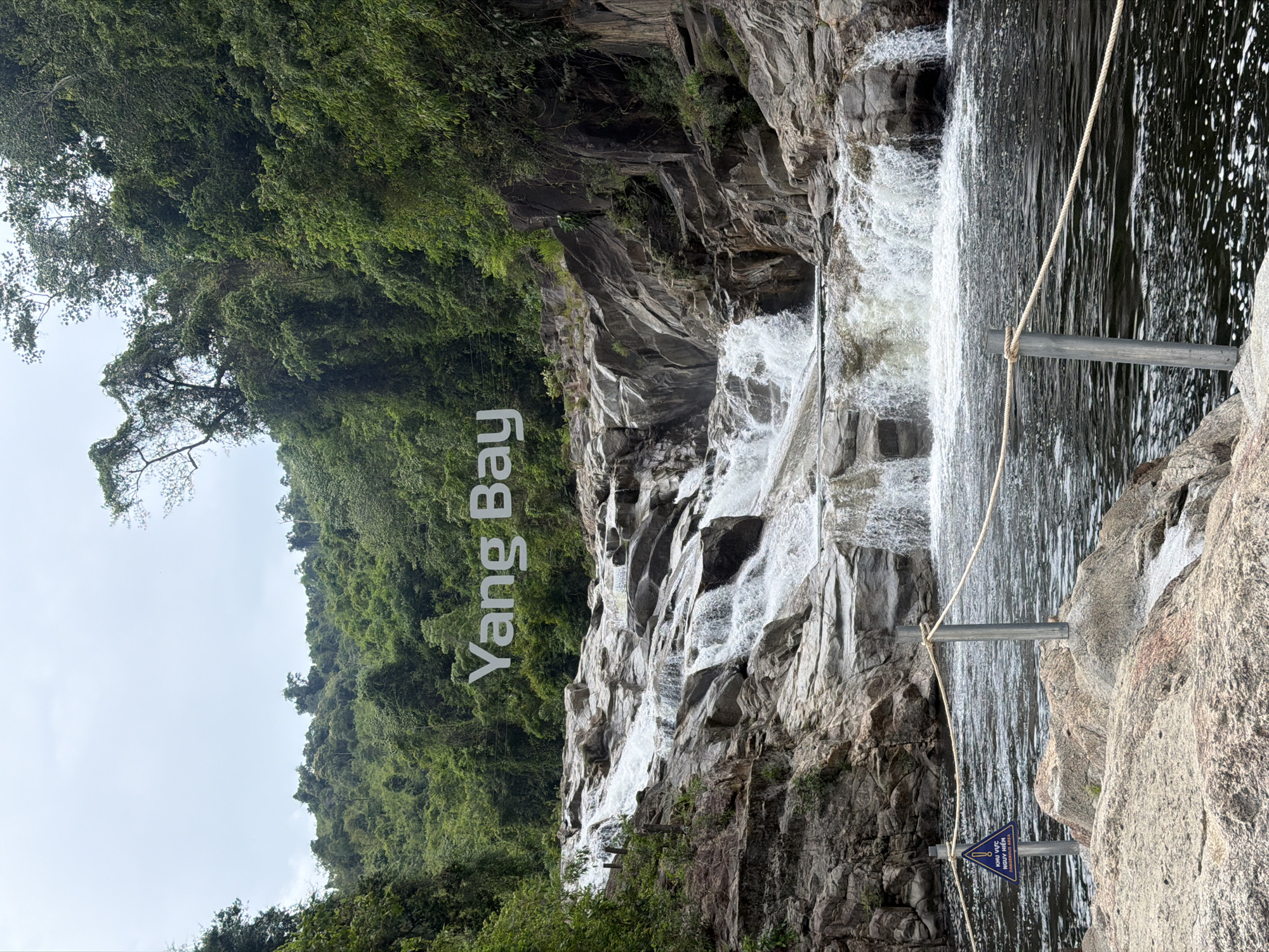 The image shows the Yang Bay Waterfall, a tourist attraction located in the city of Khanh Vinh, Vietnam. The waterfall cascades down a series of rocky terraces, with lush green vegetation covering the surrounding hillsides. The water is a deep, dark color, and a rope barrier with metal posts demarcates a safe viewing area in the foreground. A triangular blue sign with a white exclamation mark and text in Vietnamese and English indicating "Dangerous Area" is visible on the left side of the image. The sky is overcast, casting a soft light on the scene.