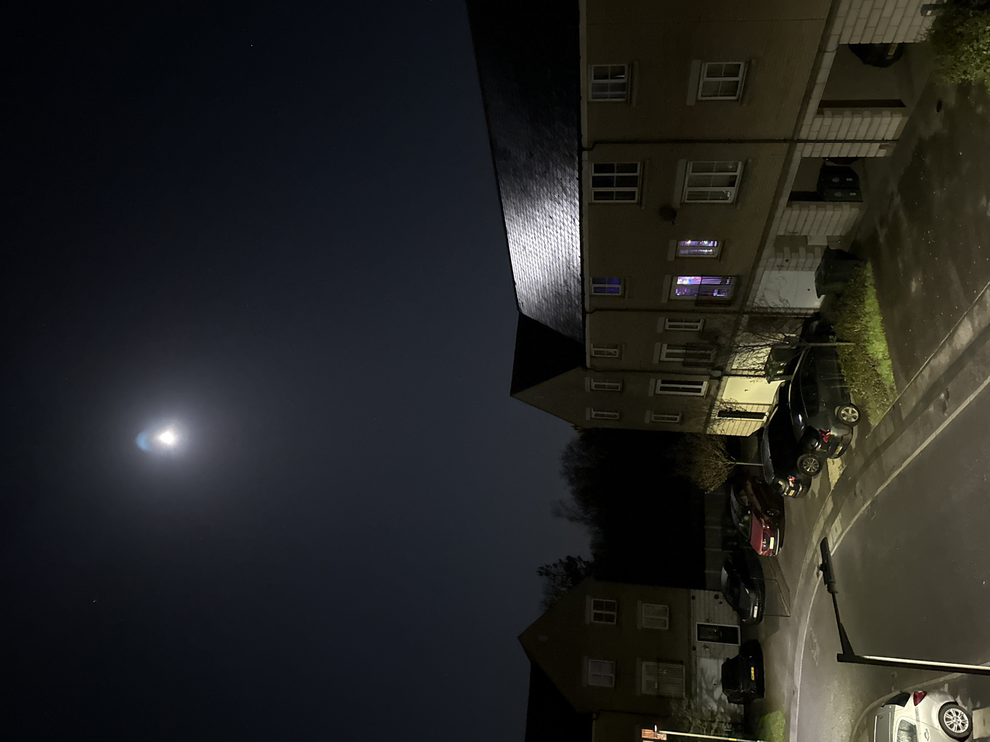 The image depicts a nighttime scene in a residential area in the city of Great Yarmouth, United Kingdom.
The sky is dark with a bright, round object visible, possibly the moon. Two residential buildings are visible. The buildings appear to be multi-story apartments with multiple windows on each level. Some windows are illuminated from within, indicating occupancy.
A curved street passes in front of the buildings. Several parked cars are visible along the street. Streetlights are present, providing some illumination to the road and sidewalk. There is also some landscaping around the building, including grass and small trees.