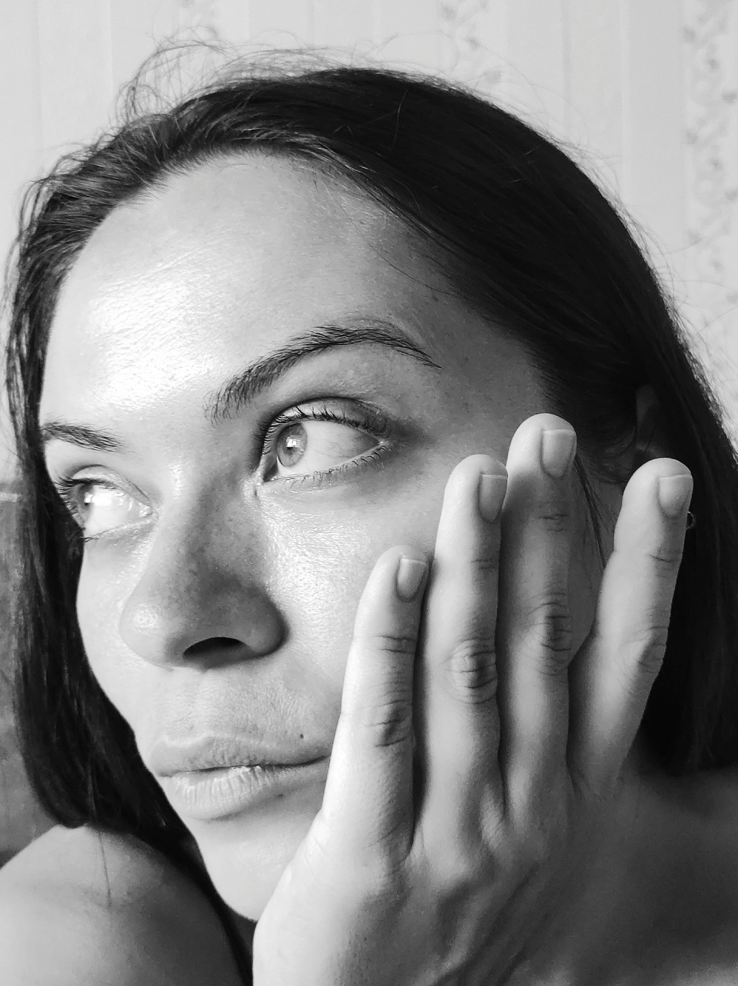 The image is a close-up, black and white portrait of a woman's face. She has dark hair and is looking to her left. Her right hand is raised, with four fingers touching her cheek and temple. The background is a textured wall, possibly wallpapered, with a subtle pattern. The lighting highlights the contours of her face, creating shadows and drawing attention to her eyes and the texture of her skin. The woman appears to be indoors. The location in Chaikovsky, Russia cannot be determined from the image.