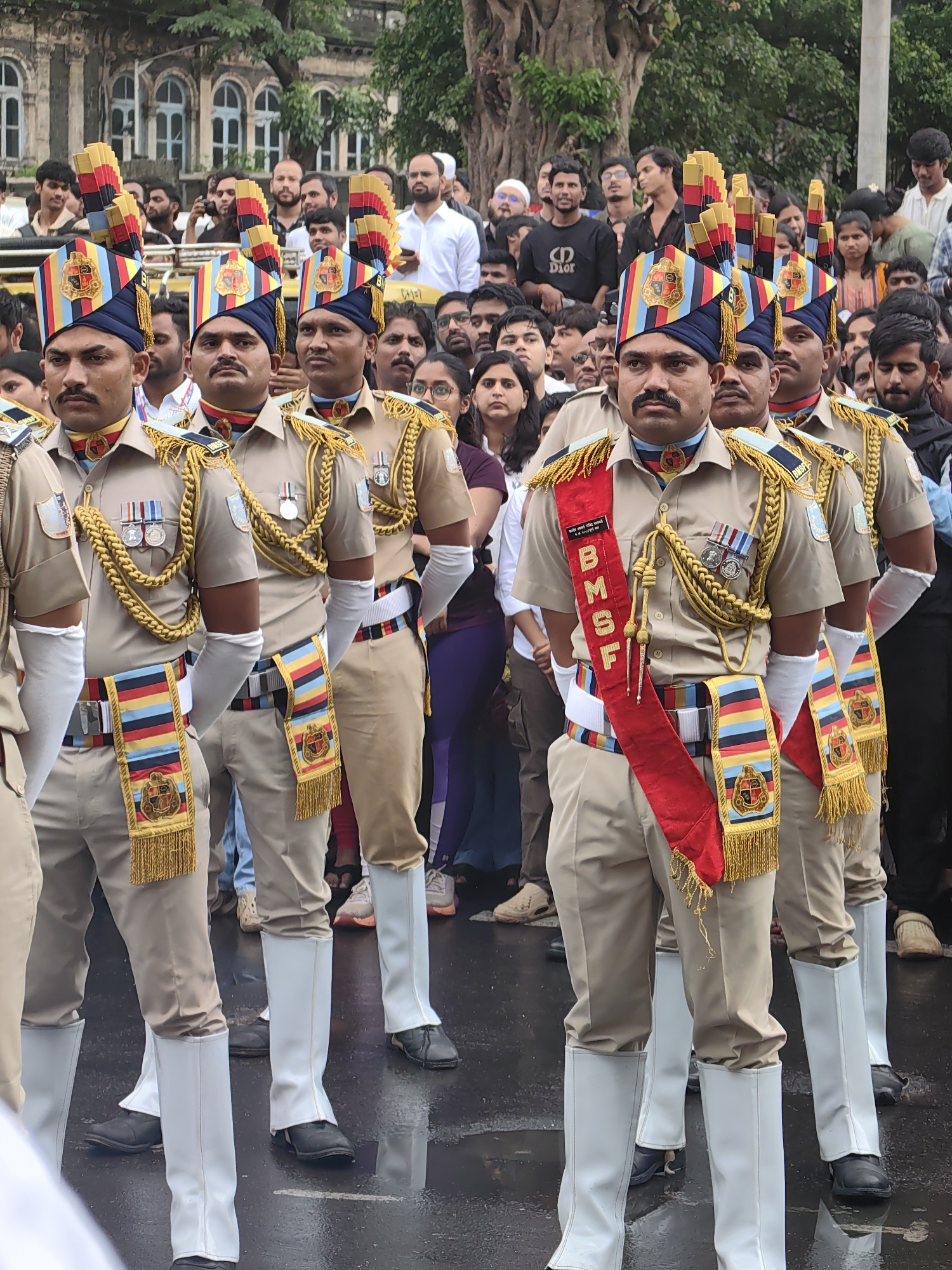 The image depicts a parade or ceremony in Mumbai, India, likely on a street with a historic building visible in the background. Several men in uniform, part of the Border Security Force (BSF), are prominently featured, standing in formation. They wear tan uniforms with elaborate gold braided epaulets, red sashes with BSF insignia, and distinctive turbans with feather-like decorations. They are also wearing white gaiters over their boots. A crowd of onlookers, dressed in civilian clothing, stands behind the uniformed individuals, observing the event. The ground appears wet, suggesting it might have recently rained or is an area prone to dampness. The presence of the BSF suggests a security or ceremonial occasion.