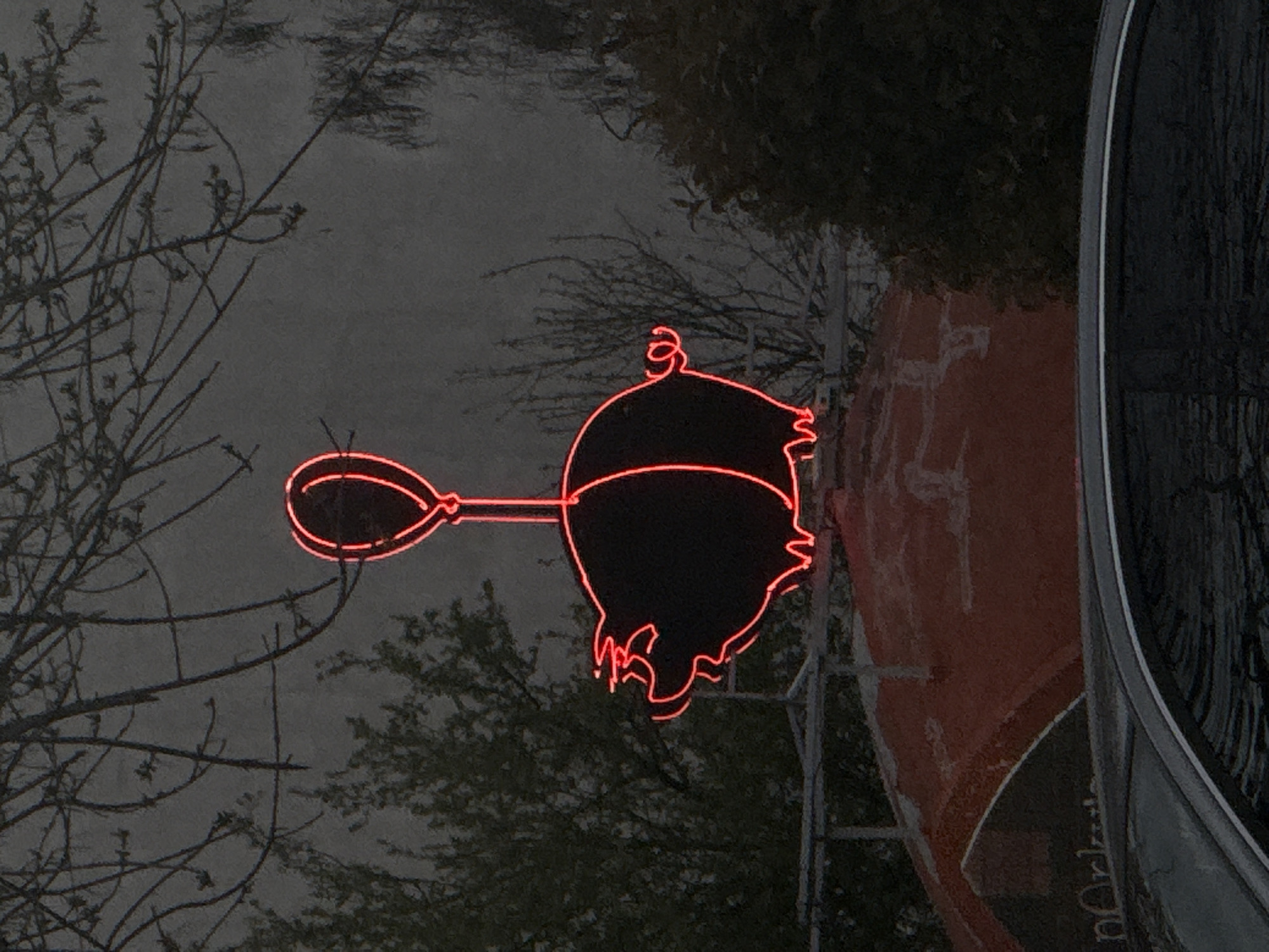 A neon sign depicting a pig holding a balloon is attached to a building's exterior wall. The wall appears to be brick or concrete. The sign is outlined in red neon. The background shows trees and a cloudy sky. Part of a vehicle is visible on the right side of the frame. A partial inscription on the building's side, next to the sign, reads "n Orle". The location is Plovdiv, Bulgaria.