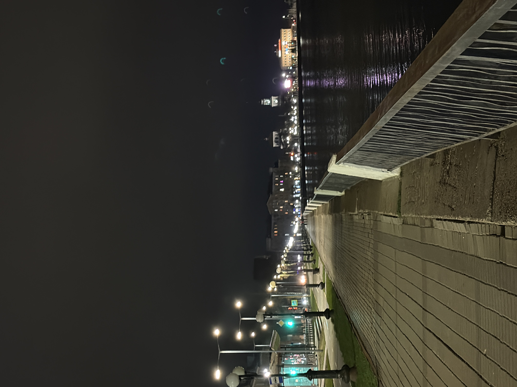 This nighttime photograph captures a serene city embankment in Votkinsk, Russia. The scene is illuminated by artificial lights, set against a dark sky. On the right, a wide, paved promenade stretches into the distance, constructed from light-colored rectangular tiles or bricks. A series of modern streetlights with multiple fixtures brightly light the walkway and a narrow strip of grass separating it from a road. A green traffic light is clearly visible further down the road, along with distant vehicle lights.
To the left of the walkway, a low, decorative railing with a top surface featuring a wavy, striped pattern borders a calm body of water, likely a river or lake. Across this dark water, the distant cityscape of Votkinsk is visible, with numerous buildings illuminated. Their bright lights create vibrant reflections on the water's surface. The dark night sky above shows some faint, crescent-shaped light artifacts, possibly lens flares. There are no people visible, suggesting a quiet moment. No discernible text is present in the image. The overall impression is one of peaceful urban tranquility.