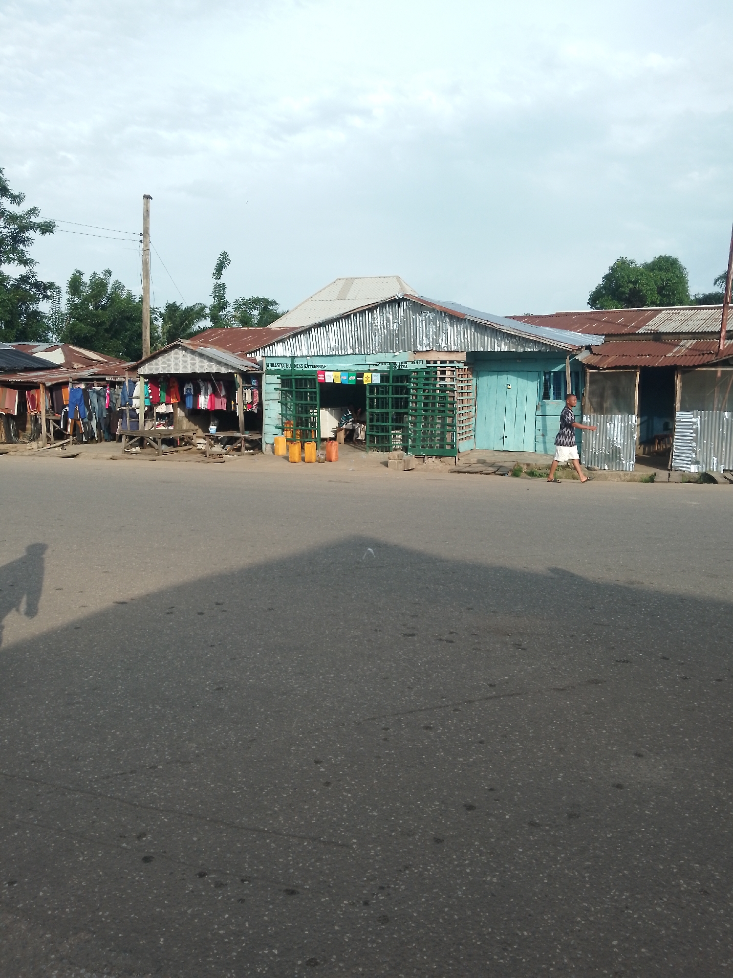 The image depicts a street scene in Serti, Nigeria. In the foreground, a paved road occupies most of the frame, with a prominent shadow of a person cast on its surface from the left. Behind the road, a row of small shops or market stalls are visible. These structures are constructed with corrugated metal and wood, and some have rusted tin roofs.
Several stalls display clothing on hangers and racks. One stall features a bright turquoise wooden facade with a green metal security gate. Yellow plastic jerry cans are placed outside this stall. A person wearing a patterned shirt and white shorts is walking from right to left in front of the shops. Further to the right, another stall appears to be more open, with corrugated metal siding.
The background shows a line of trees and a utility pole with wires. The sky above is overcast with a mix of light and dark clouds. The overall impression is of a typical rural or semi-urban marketplace in Nigeria, characterized by simple structures and the presence of goods for sale.