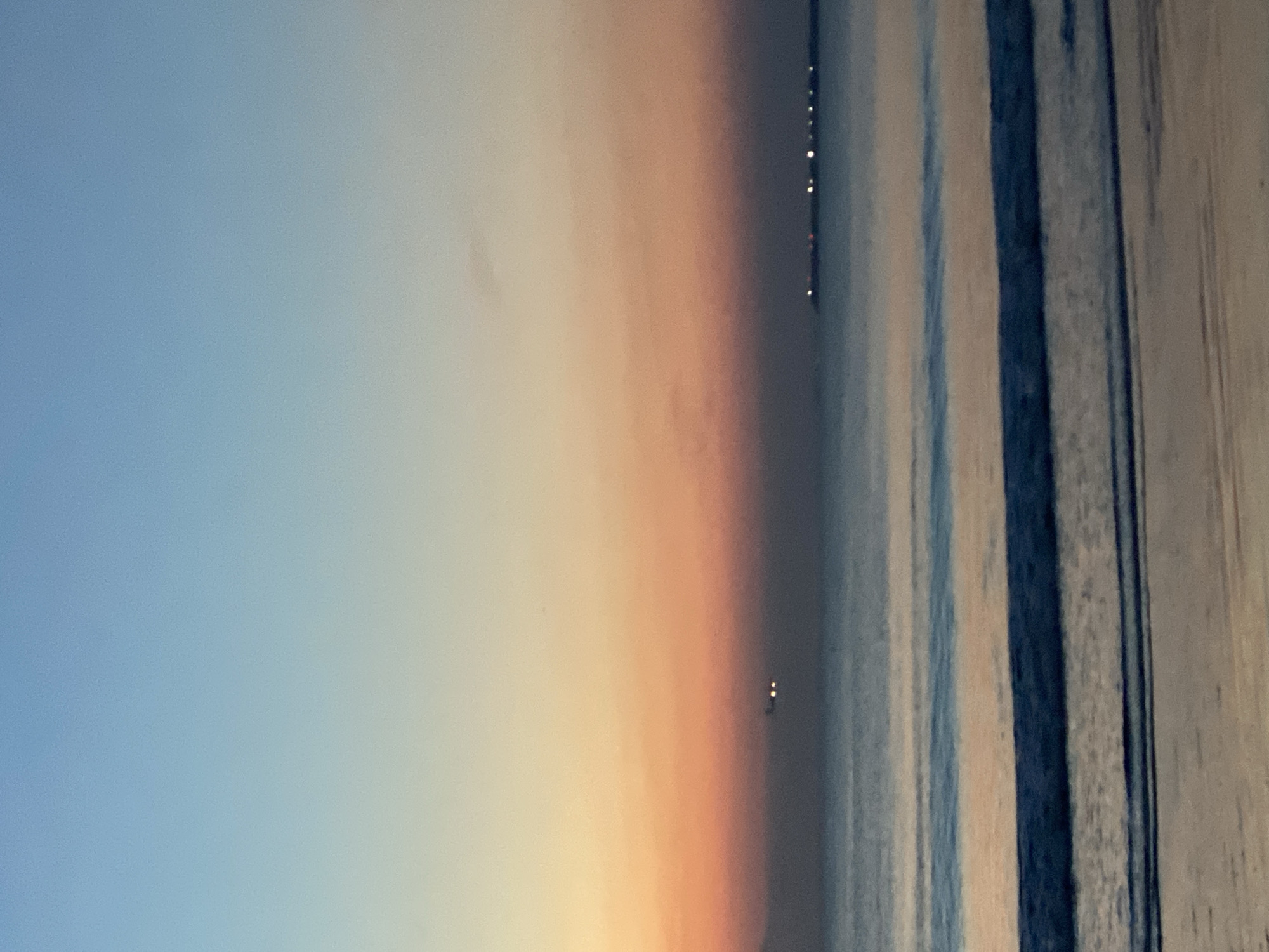 The image depicts a twilight or sunset scene. The sky transitions from blue on the left to a gradient of yellow and orange in the center. Distant lights are visible on the horizon in the middle of the image. To the right, there are vertical lines of varying shades of blue, potentially representing ocean waves or an object with a striped pattern. The location is possibly Kuta Selatan, Indonesia, as lights from land are seen on the horizon.