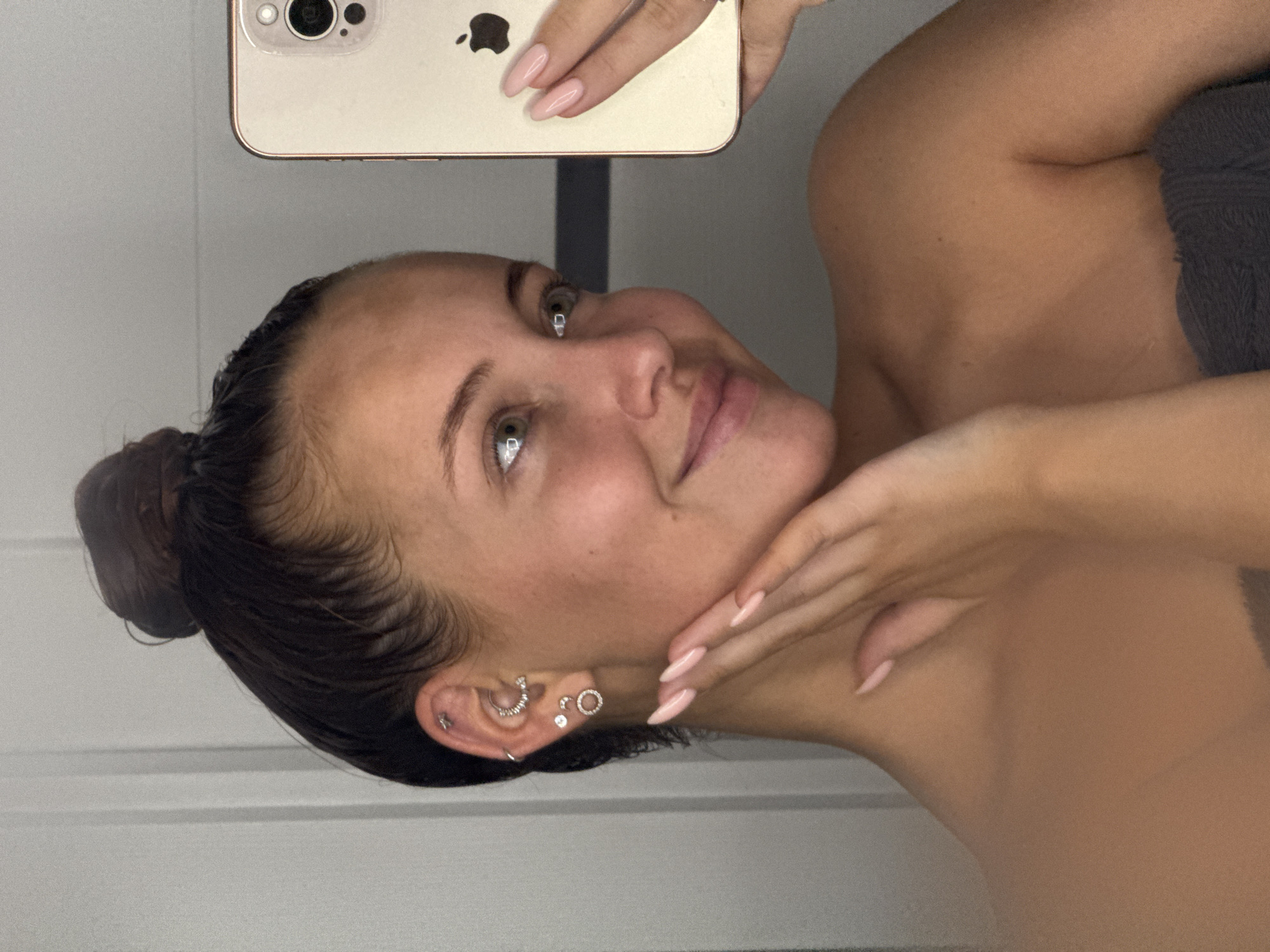 A young woman with dark hair pulled into a topknot is taking a selfie in a mirror. She is wearing a grey towel around her shoulders. Her left ear is adorned with multiple piercings, including studs and a hoop. She is touching her face with her right hand, which has neatly manicured pale pink nails. The background appears to be a bathroom with white vertical panels and a grey wall. The woman's expression is a slight smile with her eyes looking off to the side. The reflection of an iPhone is visible in the mirror. The scene does not provide any specific geographical indicators to pinpoint a location in Chaikovsky, Russia.