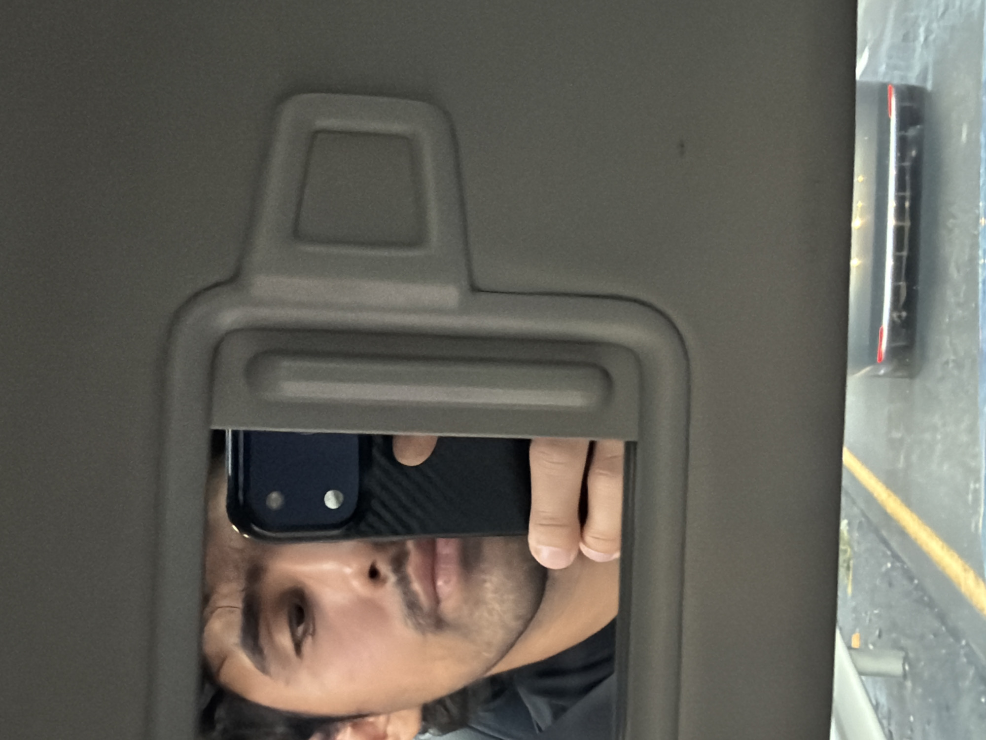 A selfie taken in the mirror of a car's sun visor. The person's face, hand and phone are reflected in the mirror. A car door is visible outside the car window. The location in San Bernardino Chalchihuapan, Mexico, cannot be determined from the image.
