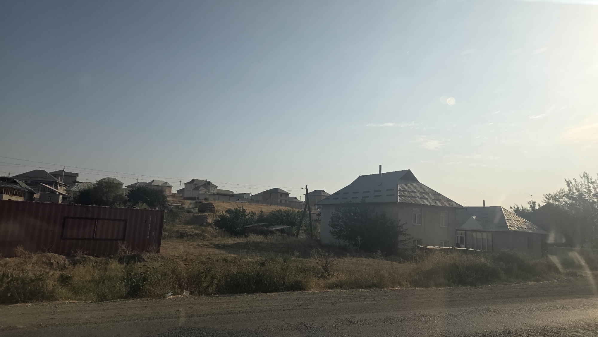 The image captures a sunlit, rural scene in Baş-Bulak, Kyrgyzstan. A row of houses, varying in architectural style and size, are situated on a gently sloping hill. The houses feature pitched roofs, some with visible chimneys. In the foreground, a large, weathered, reddish-brown shipping container is positioned on a dry, grassy embankment. Beyond the container and the houses, the sky is a pale blue with a hazy, bright sun illuminating the upper right portion of the frame, creating a lens flare effect. The ground in the immediate foreground appears to be a dirt or gravel road. The overall impression is one of a developing residential area on the outskirts of a town, characterized by simple structures and dry, natural vegetation. There are no people visible in the image.