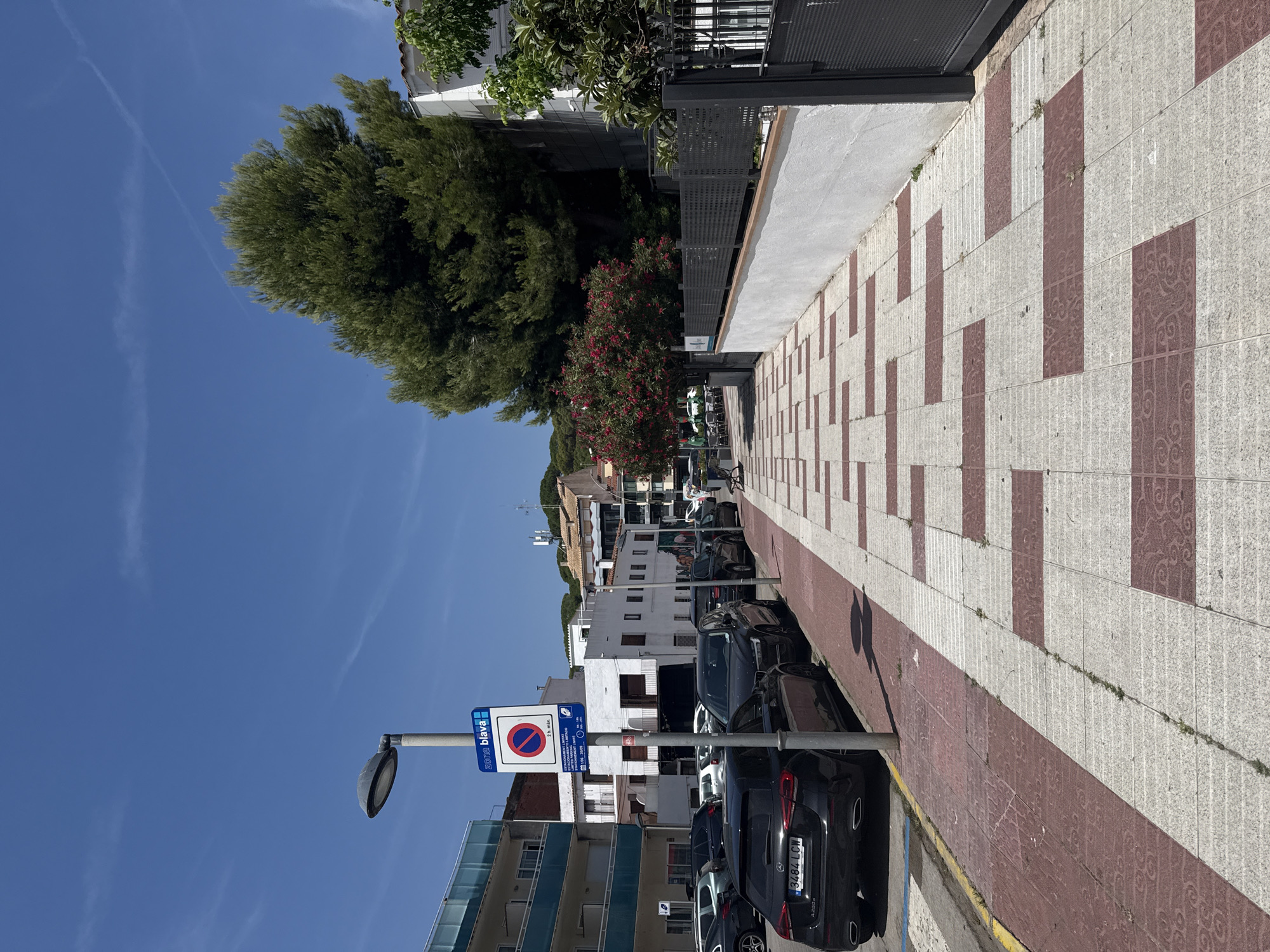 The image depicts a street scene in Platja d'Aro, within the municipality of Castell d'Aro i s'Agaró, Spain, under a clear blue sky with thin white clouds. The foreground features a sidewalk with a repeating pattern of white and red rectangular tiles, some of which have decorative swirling motifs. Along the left side of the sidewalk, cars are parked, including a black Mercedes-Benz A-Class with a license plate reading "3484 LCW". A blue and white parking sign with the text "ZONA blava" and a prohibition symbol indicating "2 h. max." is mounted on a lamppost. Beyond the parked cars, buildings with balconies and windows are visible, with a large pine tree dominating the right side of the frame, obscuring part of a white building. A grey metal fence runs along the right edge of the sidewalk. In the distance, more buildings and trees can be seen. The scene is devoid of any visible people.