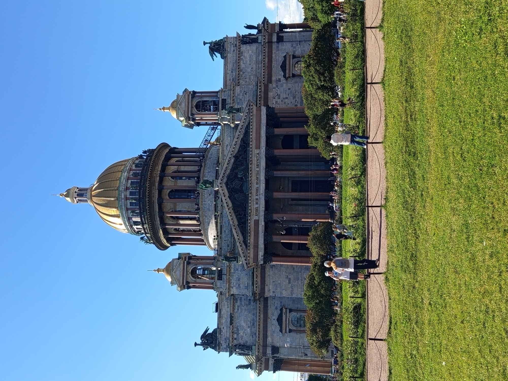 The image shows a wide-angle view of a grand, historic building with a golden dome, likely St. Isaac's Cathedral in Saint Petersburg, Russia. The building is constructed of stone and features multiple levels with columns, statues, and architectural details. The sky is a clear, bright blue, indicating a sunny day.
In the foreground, a well-maintained grassy lawn is visible, with a paved walkway running alongside it. Several people are present, some walking on the path and others sitting or standing on the grass. The people are casually dressed, suggesting a leisurely outing. Two individuals in the foreground are looking at something on the path, possibly a map or brochure. One person is wearing a white t-shirt and blue jeans, and the other is wearing a white top and a light-colored head covering.
The time of day appears to be afternoon, given the bright sunlight casting shadows. The overall atmosphere seems peaceful and relaxed, with people enjoying the architecture and the outdoor space. The image captures a blend of historical grandeur and everyday life. No specific text is clearly legible in the image.