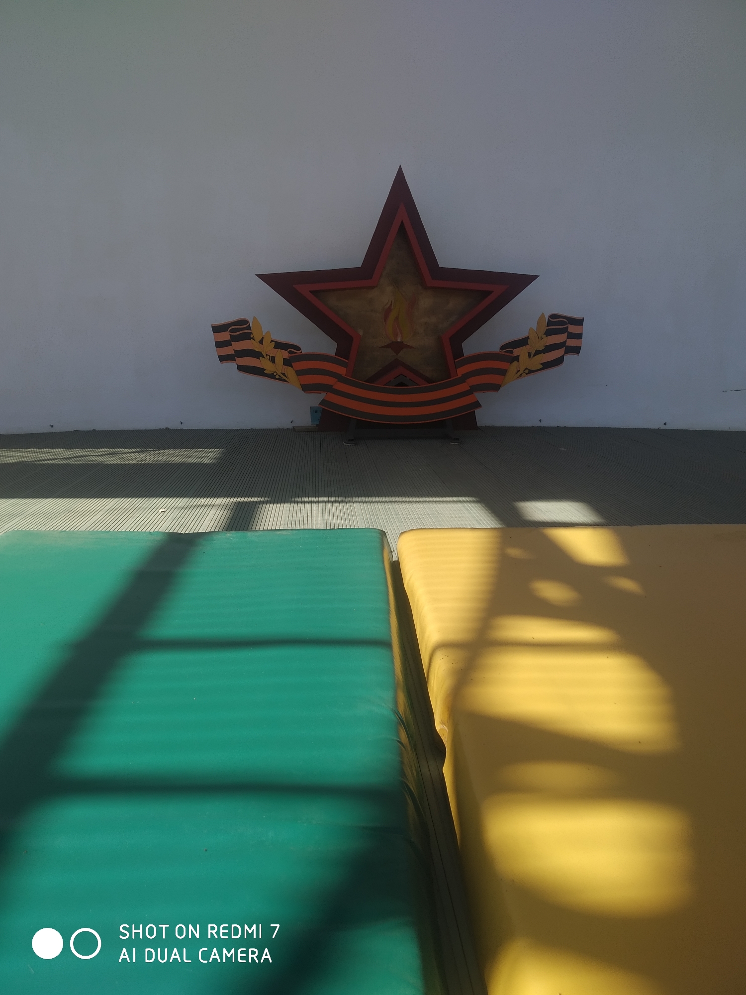 The image shows a stylized red star, a symbol often associated with the Soviet Union and Victory Day, mounted on a white wall. The star is outlined in a darker red or maroon and contains a depiction of a burning flame within. A black and orange ribbon, reminiscent of the Ribbon of Saint George, flanks the star on both sides, with decorative laurel leaves incorporated into the design.
The scene appears to be indoors, with the star mounted on a wall above a floor with a patterned surface. In the foreground, there are two padded surfaces, one green and one yellow, positioned side-by-side. The lighting suggests sunlight streaming in from an unseen source, casting shadows across the floor and the padded surfaces.
While the exact location within Chaikovsky, Russia, cannot be definitively determined from the image alone, the presence of the stylized red star and the Ribbon of Saint George strongly suggest a location that commemorates Soviet-era history or Victory Day celebrations.