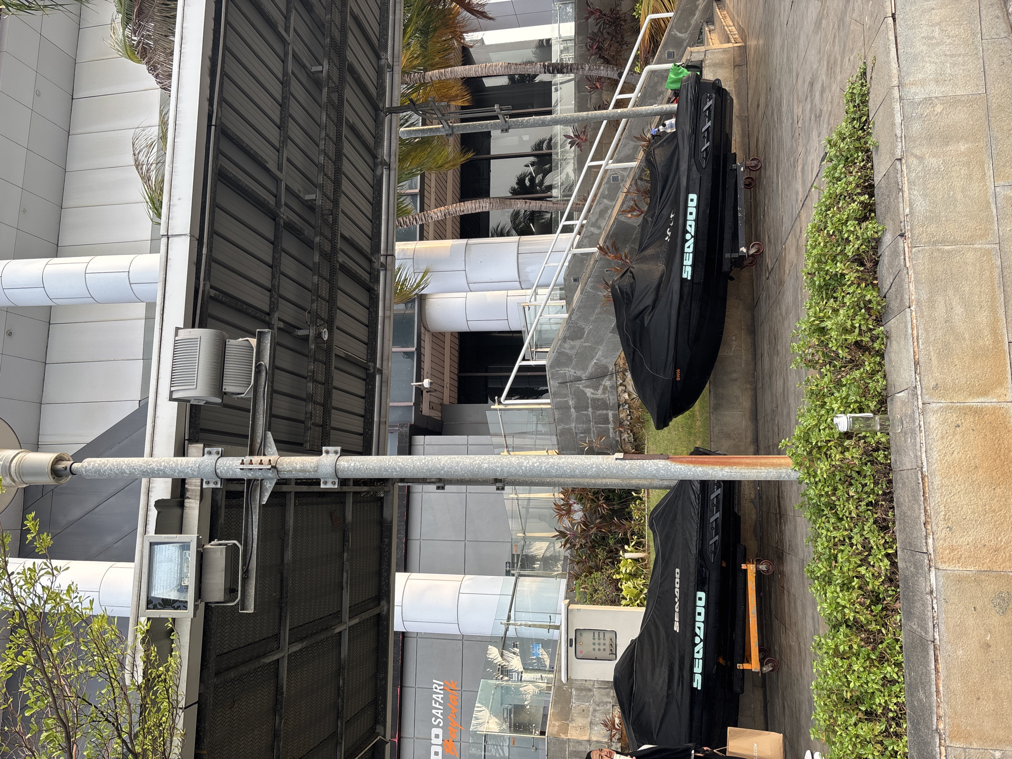 Two jet skis covered in black tarps are parked on a paved walkway next to a modern building with glass windows and dark metal siding. A white staircase with metal railings leads up beside the jet skis. Lush green plants grow along the edge of the walkway and between the jet skis and the building. One of the jet skis has "SEA-DOO" written in a teal-colored font on its cover. A green bottle rests on the railing above the first jet ski. The scene appears to be an outdoor area in front of a commercial or residential building, possibly a marina or a waterfront property, given the presence of jet skis. The lighting suggests it is daytime, and the weather seems clear. In the background, palm trees are visible, and their reflections can be seen in the building's windows, hinting at a tropical or subtropical climate. There is also a sign that says "SAFARI" on the building.