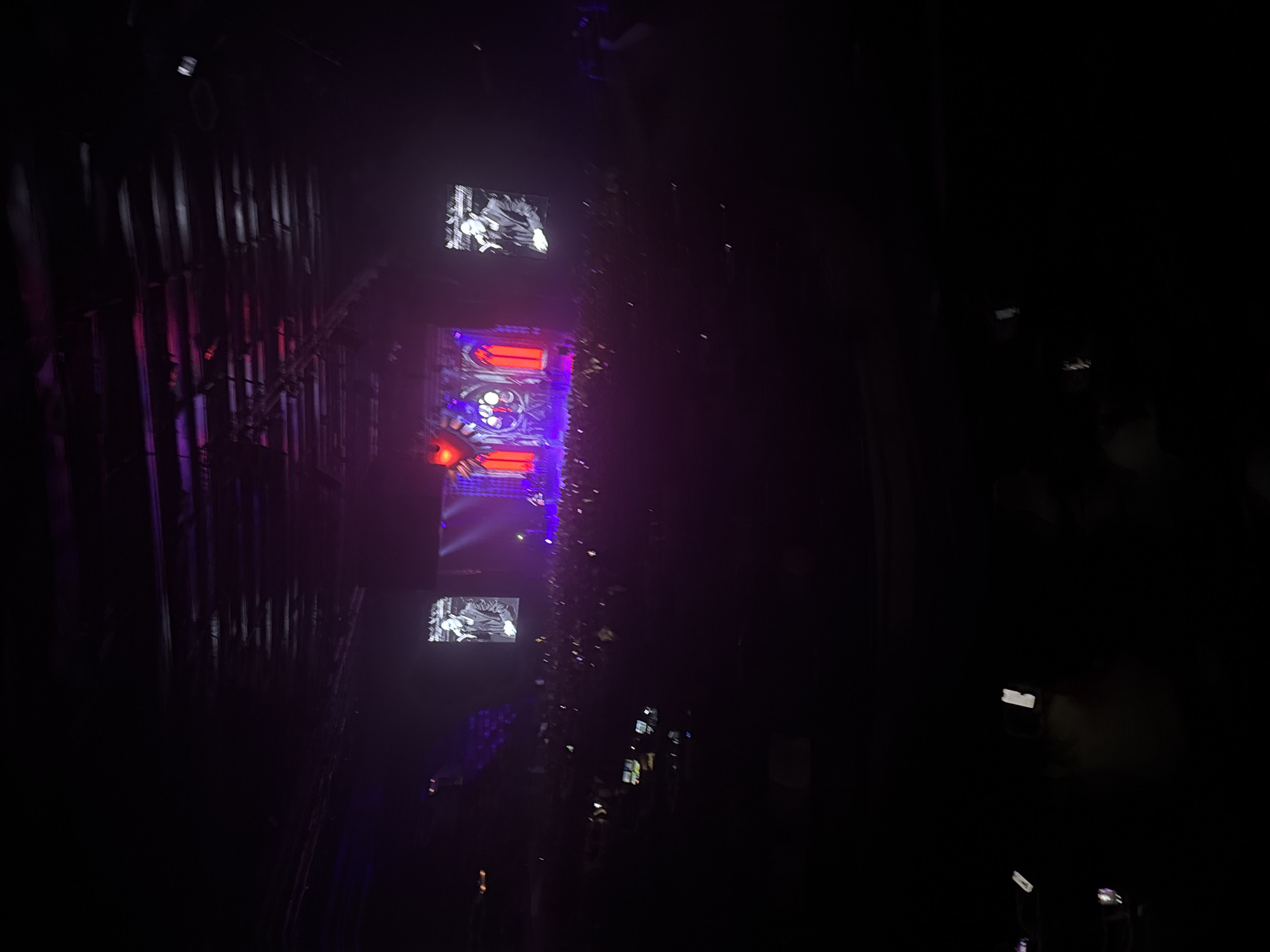 The image depicts a nighttime scene in what appears to be a concert or performance venue, possibly in Perm, Russia, given the prompt's reference. The central focus is a vertically-oriented structure featuring multiple screens displaying visual content. The top and bottom screens each show black-and-white imagery, possibly a performance clip or abstract video art. The middle screen displays a complex, brightly illuminated, gothic-style design with a mix of red and blue/purple lighting. The structure is flanked by what look like metal pipes or scaffolding on the left. Light fixtures and possibly decorative foliage adorn the sides of the structure. The overall environment is dark, suggesting the venue is dimly lit or the lighting is concentrated on the structure itself. In the far distance, some blurry indistinct shapes are present which may be other buildings or structures.