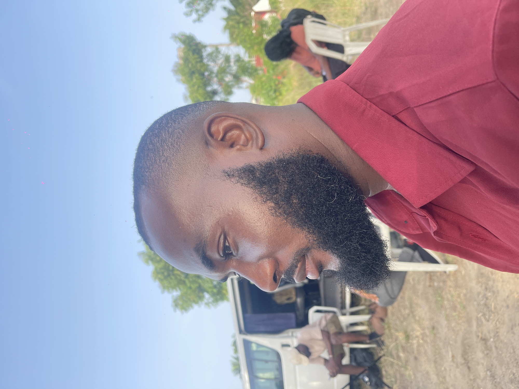 A medium shot depicts a man with a shaved head and a full, dark beard, wearing a burgundy shirt. He appears to be outdoors during daylight. The background is blurred but shows a light-colored van, other individuals, and possibly trees. The location appears to be Unknown, Unknown.