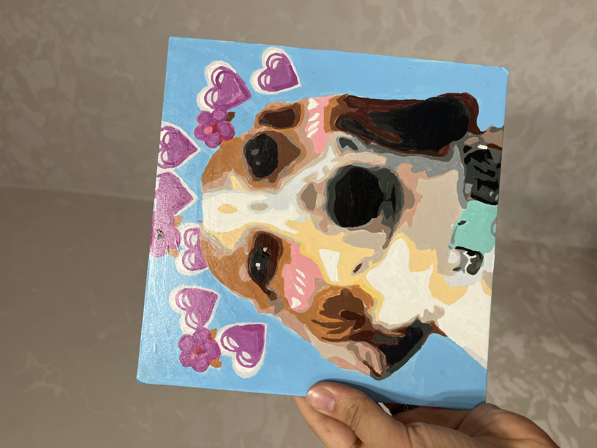 A square painting depicting a dog's head surrounded by hearts and flowers is being held by a hand against a neutral background. The painting uses distinct blocks of color to create the image, with a light blue background. The dog has brown, white, and black patches. Purple hearts and pink flowers are scattered around the dog's head. The hand holding the painting has light skin and a short fingernail. The background appears to be a neutral-colored wall.