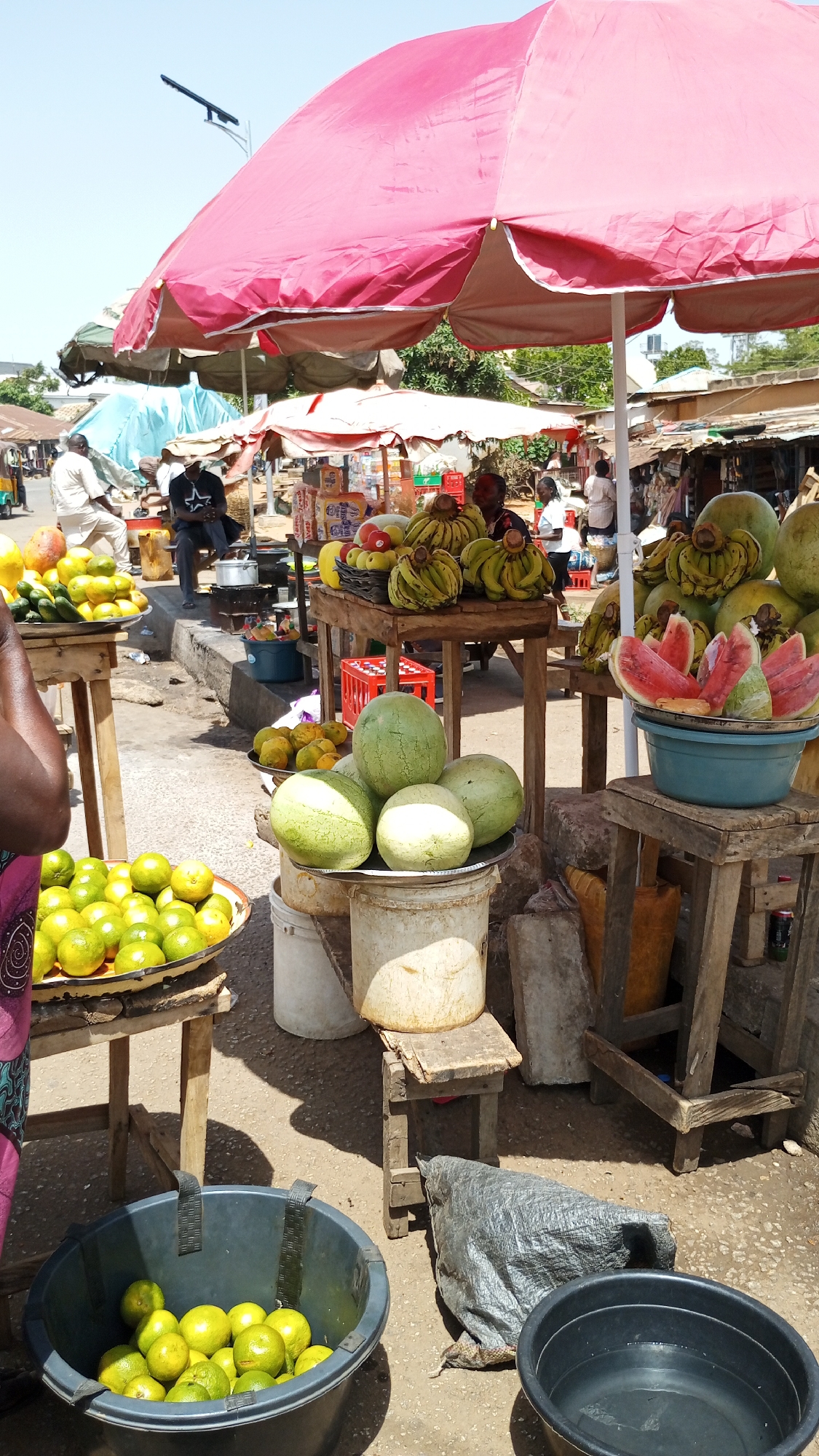 The image shows a fruit stand in Bukuru, Nigeria. Underneath a large, red umbrella, various fruits are displayed on wooden tables and in buckets: mangoes, bananas, oranges, watermelons, and unidentified green fruit. People are visible in the background, including individuals seated and standing near other stalls or vehicles. The environment is urban, with buildings and streets evident in the background. Containers and other support structures are placed underneath the tables holding the fruit, and the ground appears to be a mix of pavement and dirt.