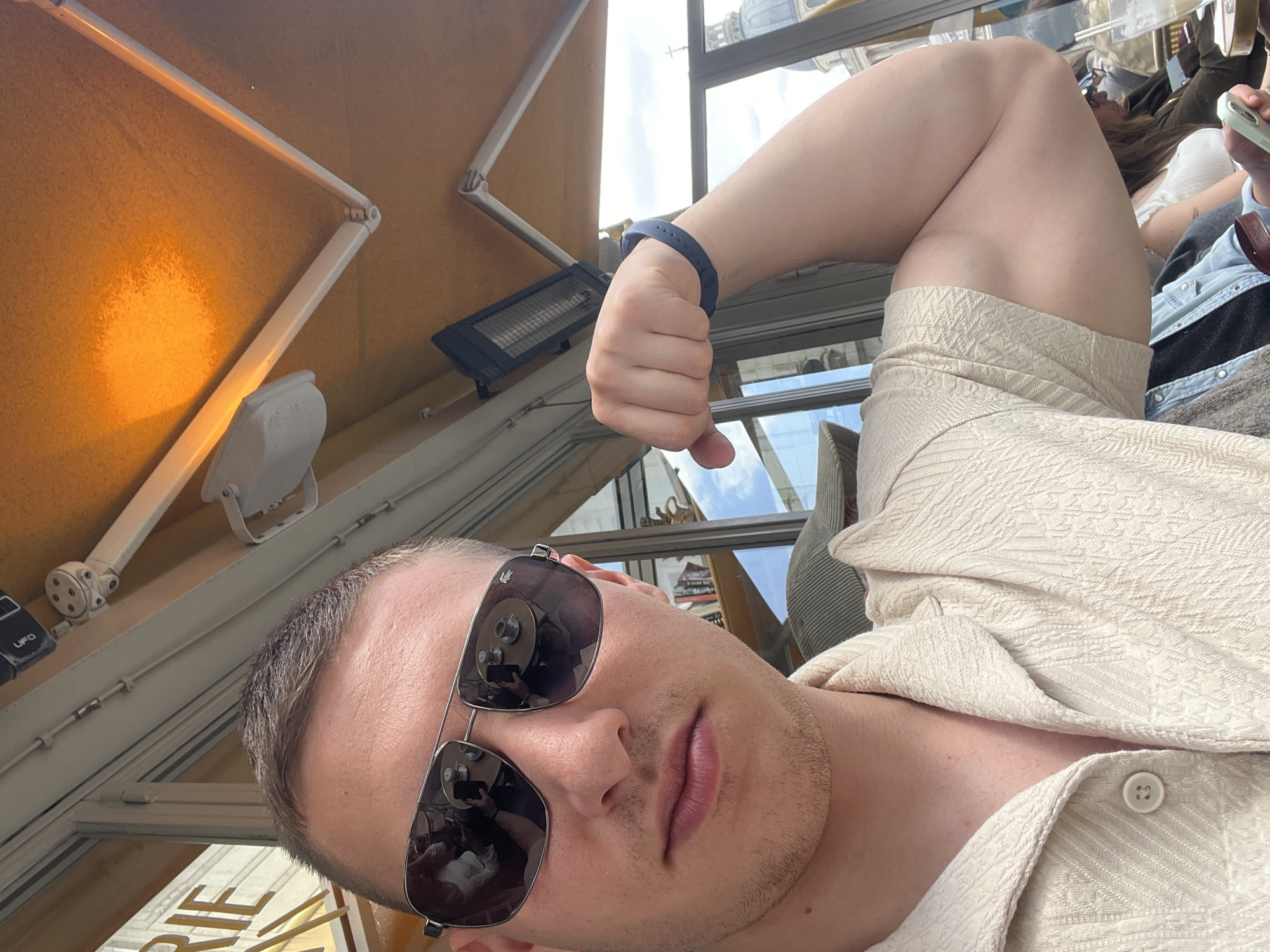 The image is a tilted, close-up selfie of a man, taken from a low angle. He has short, dark hair and light stubble, and wears silver-framed aviator-style sunglasses that reflect the surroundings. His lips are slightly parted. He is dressed in a light beige or cream-colored short-sleeved shirt with a textured, patterned fabric and visible buttons. On his left wrist, he wears a dark blue fitness tracker or bracelet. His left arm is bent, with his hand in a clenched fist and his thumb pointing downwards.
The setting appears to be an outdoor terrace or balcony, enclosed by glass panels, suggesting a café or restaurant. Overhead, white structural elements, a spotlight, and a dark outdoor heater are visible against a warm, possibly yellow, ceiling or awning. Through the glass and in the sunglasses' reflection, bright daylight and a blue sky are seen. A large, metallic dome, characteristic of Parisian architecture, is discernible in the background, confirming the location in Paris, France.
Other people are partially visible in the background: one person in the upper right holds a light green phone, wearing a light top and a denim jacket. Another person's leg, clad in a light-colored patterned skirt or dress, is seen in one of the crops, indicating they are seated nearby. Visible text includes "UFO" on a black device overhead and a partial word, "RIE", likely part of a French shop name, on a sign through the glass. The bright lighting suggests it is daytime. The man's expression is obscured by the sunglasses, but his pose appears casual.