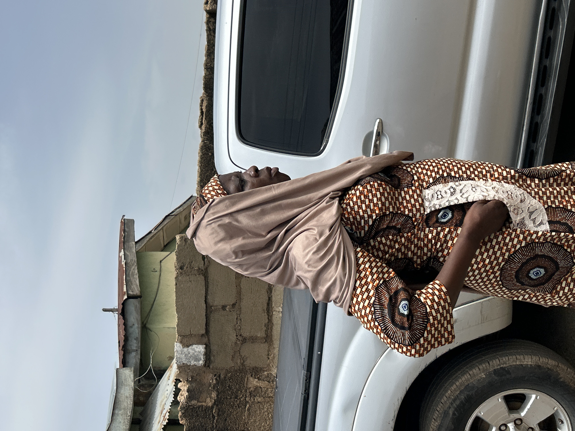A woman lies across the hood of a silver truck. She is wearing a patterned brown dress with white lace and a light brown head covering. The truck's tire is visible in the foreground. In the background, there is a building with a partially collapsed roof. The sky is overcast. The location is in a built environment, likely a neighborhood in Jos, Nigeria, given the context.