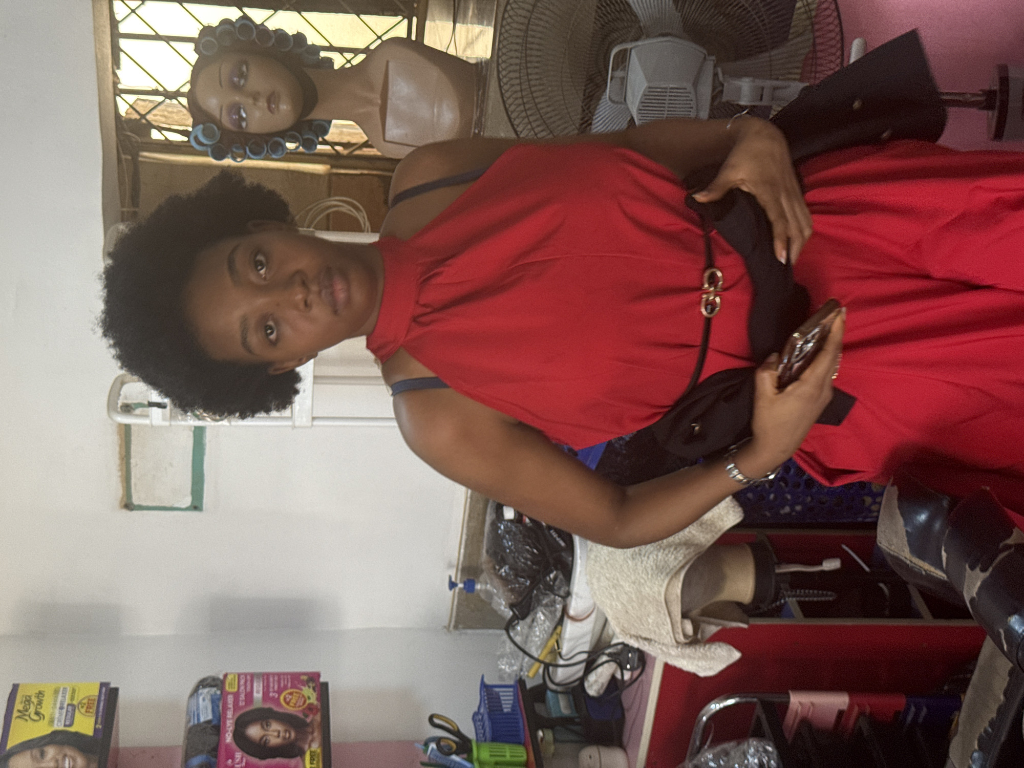 A woman in a red dress with a black belt is holding a cell phone in a room in Abuja, Nigeria. She is standing near a table with hair products and accessories, including boxes of hair relaxer, a mannequin head with hair rollers, and a fan. The room appears to be a hair salon or a personal grooming space. The light entering through the window suggests a daytime setting.