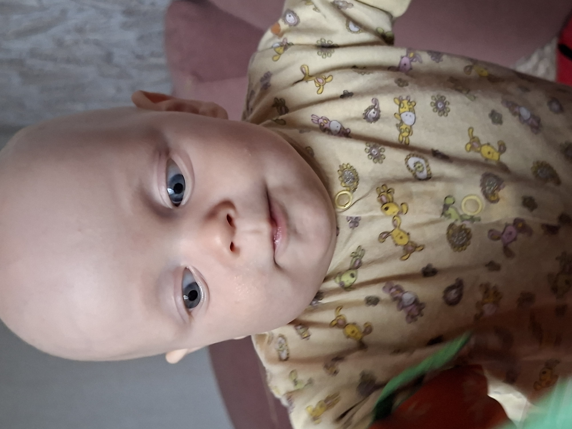 The image is a close-up, vertically oriented photograph of a baby. The baby has fair skin and light blue eyes. Their head is tilted to the side, and they are wearing a pale yellow onesie with a pattern of cartoon animals, including giraffes and rabbits, in various colors such as yellow, pink, and purple. The onesie has snap closures, one of which is clearly visible near the baby's chest. The background is out of focus, showing what appears to be a textured surface, possibly a blanket or bedding, in a light color, and a hint of a pink soft surface, likely a piece of furniture. The lighting suggests an indoor setting, with no clear indication of the time of day. The baby's expression is neutral, and their gaze is directed slightly away from the camera. The overall impression is an intimate and candid snapshot of a young child. The provided crops show closer details of the baby's face, specifically their eye and mouth area, and the pattern on their clothing. There is no visible text in the image. The location context is Karsha, Russia.