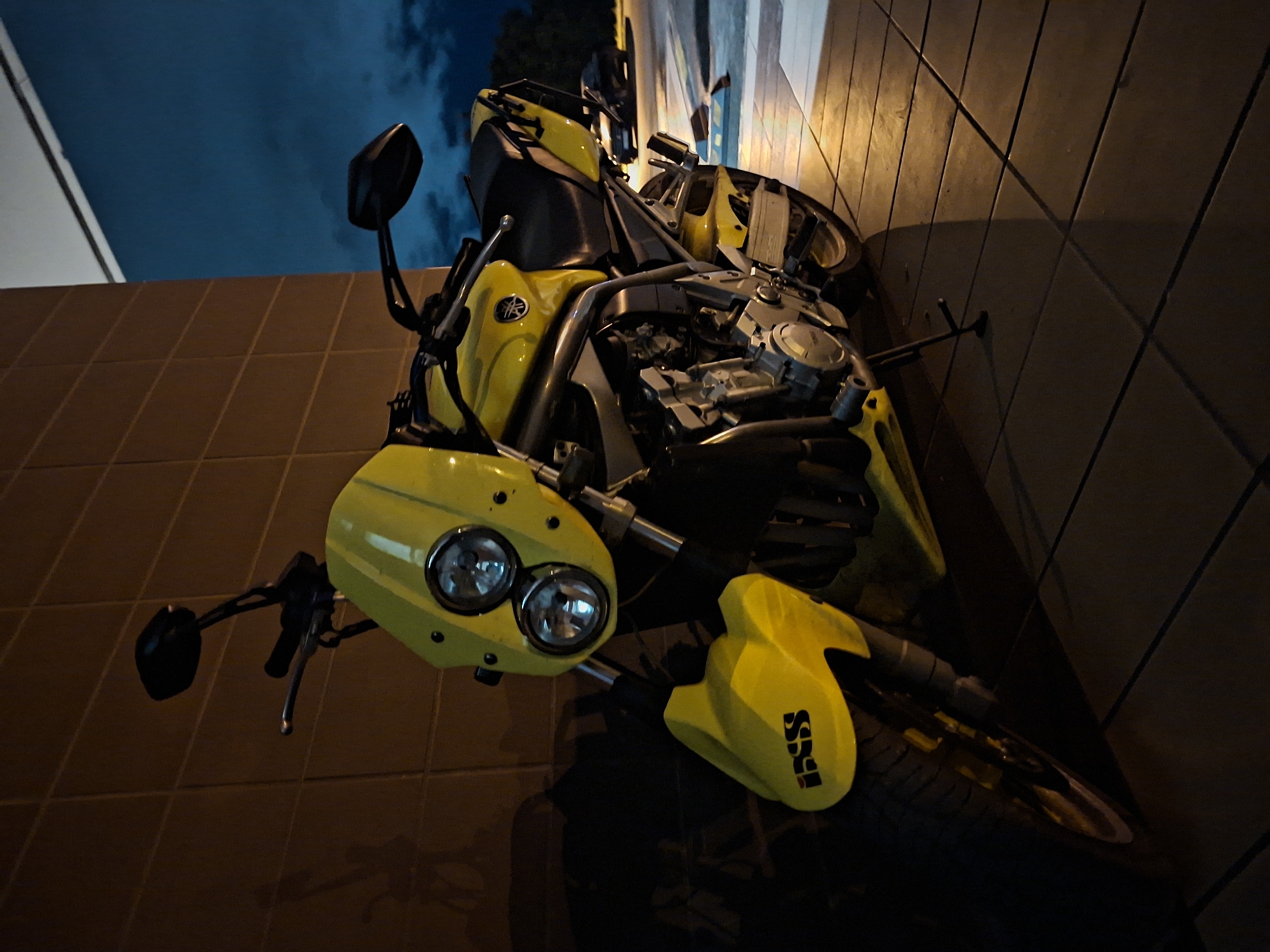 A yellow Yamaha motorcycle is parked on a tiled sidewalk at dusk. The motorcycle is angled towards the camera, with its front wheel closest. The motorcycle has dual headlights and a black seat. The background shows a brown brick wall and a dark blue sky with clouds. In the distance, another vehicle is visible parked on a paved area. The scene appears to be located in an outdoor urban setting.