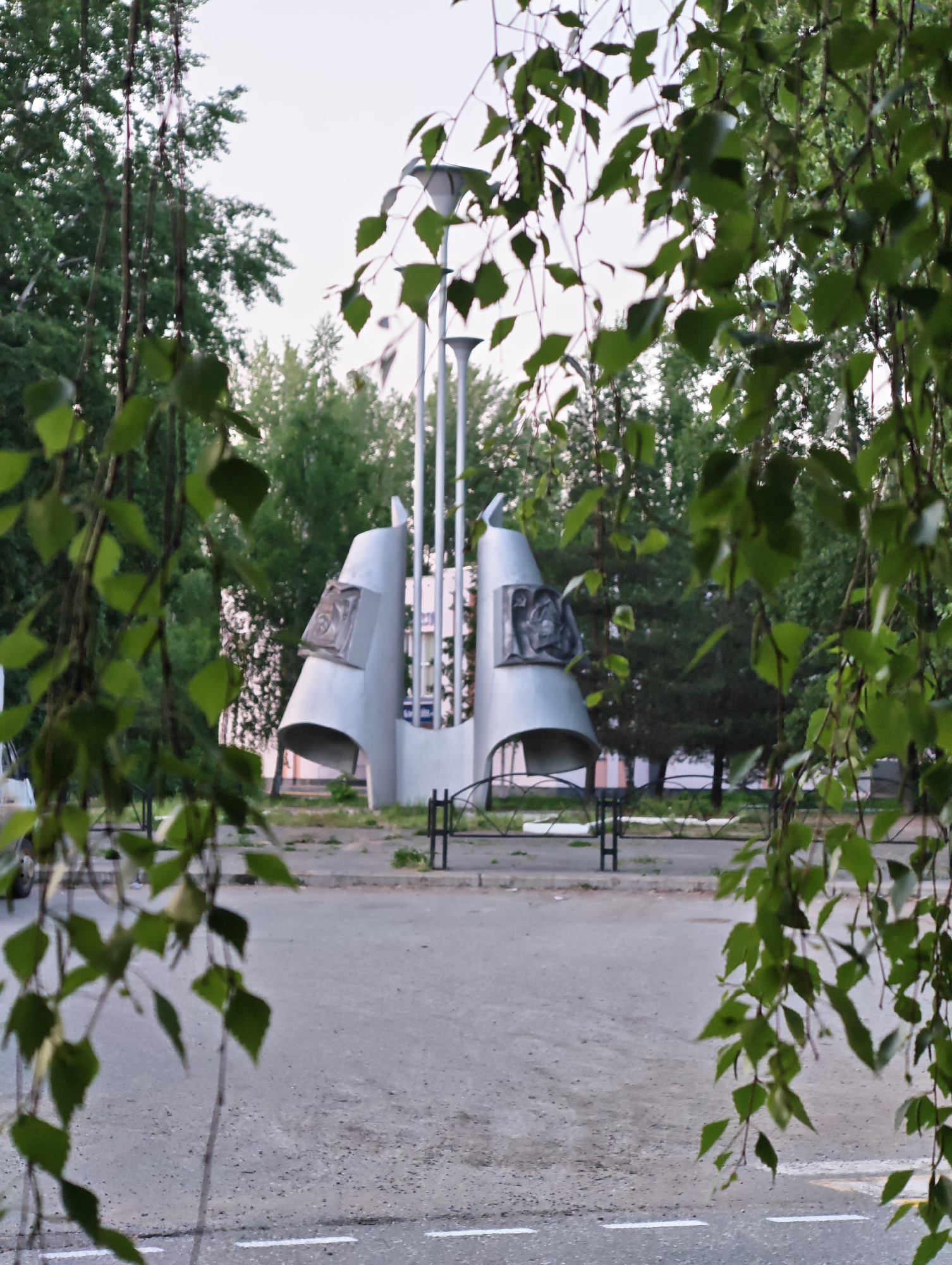 The image captures a view of the monument "Stele in honor of the founders of the city of Chaikovsky" in Russia. The monument is made of metal and has a futuristic design with vertical posts and circular elements. It is located in a public space with trees and greenery. A street runs in front of the monument, marked with white lines. Leaves from a tree in the foreground frame the scene, adding a natural element to the urban environment.