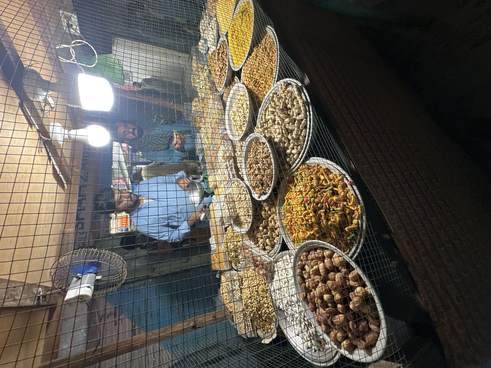 The image captures a vibrant display of various dry fruits and snacks, viewed through a dark, square-grid metal mesh. The scene appears to be an indoor or covered market stall, brightly illuminated by two prominent white LED bulbs hanging within the space.
In the foreground, the metal mesh acts as a barrier, behind which numerous round, shallow trays are arranged, showcasing an array of edible goods. These include large quantities of unshelled peanuts, shelled peanuts, dates, what appear to be yellow lentils or split peas, and a colorful mixed savory snack (possibly 'namkeen' or 'chanachur'). Other trays hold various light-colored nuts or seeds and different varieties of dried goods.
Behind the food display, two men are visible. One man, wearing a light blue shirt, is positioned centrally, looking towards the camera. Another man, also in a light blue shirt, is partially visible further back. Their presence suggests they are vendors or patrons of the stall.
To the left, a small, caged fan is mounted, and behind it, a wooden wall or shelf structure is visible. The bright artificial lighting suggests it is either night time or an interior space with limited natural light. The overall atmosphere points to a bustling market setting, common in many parts of the world for selling such snacks and dry fruits. No clearly legible text is visible within the frame.