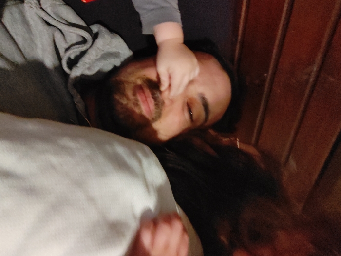 A close-up image depicts three individuals engaged in an indoor, intimate setting. In the foreground, an adult male with dark hair, a beard, and a slight smile on his face is visible, wearing a grey hooded sweatshirt. A small, light-skinned hand, clad in a grey long-sleeved garment, is playfully pressed against the male's nose and upper facial area. To the right and behind the male, a portion of dark hair and a light-colored, possibly white or cream, fabric (appearing to be clothing or a blanket) are visible, indicating the presence of a third, largely obscured individual. The background consists of dark brown, horizontally oriented wooden paneling under dim lighting. The interaction between the male and the child's hand appears playful. The location in Tel Aviv-Yafo, Israel, cannot be determined from the provided visual information.