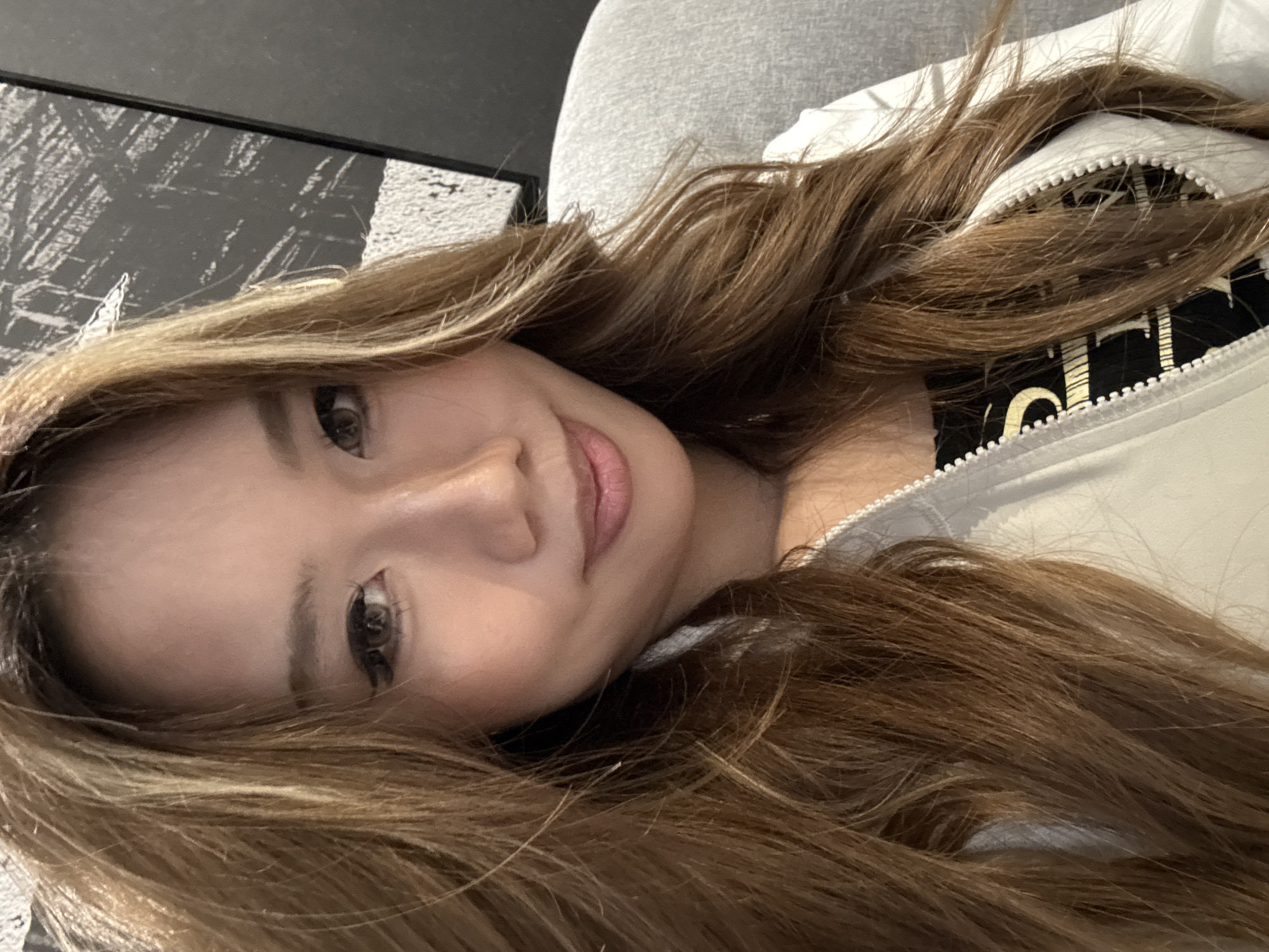 A selfie image shows a woman with long, highlighted brown hair. She is wearing a white jacket with a visible black garment underneath. The background includes a grey surface and a black and white textured area. The location is not identifiable as being in Liangjiang, China, or any other specific place.