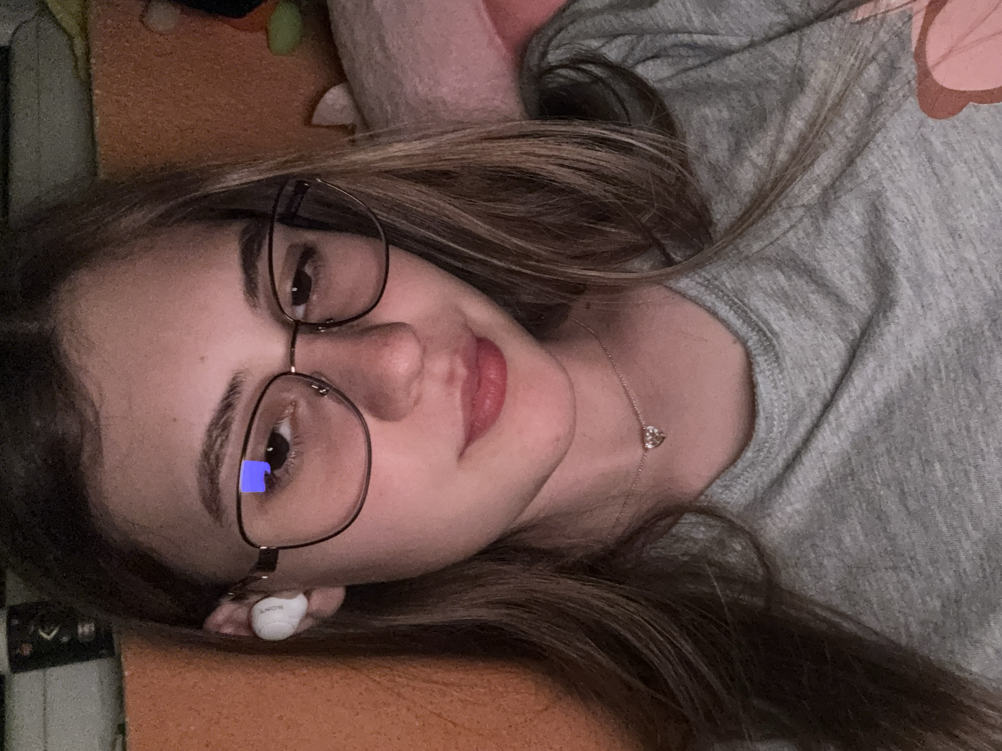 A young woman is pictured lying down indoors in Panagyurishte, Bulgaria. She has long, light brown hair spread around her and wears dark-framed glasses. Her left ear features a white wireless earbud with "SONY" visible on its side. Her brown eyes show a distinct blue rectangular reflection in the left eye, indicating light from a screen. She has a subtle pink lip color and a gentle, relaxed expression. A delicate silver chain with a small, sparkly, heart-shaped pendant rests on her neck. She is wearing a grey marled top. The background consists of soft, textured fabrics in shades of orange-brown and light pink, suggesting a comfortable setting like a bed or couch. The lighting is soft, characteristic of an indoor environment, possibly in the evening.