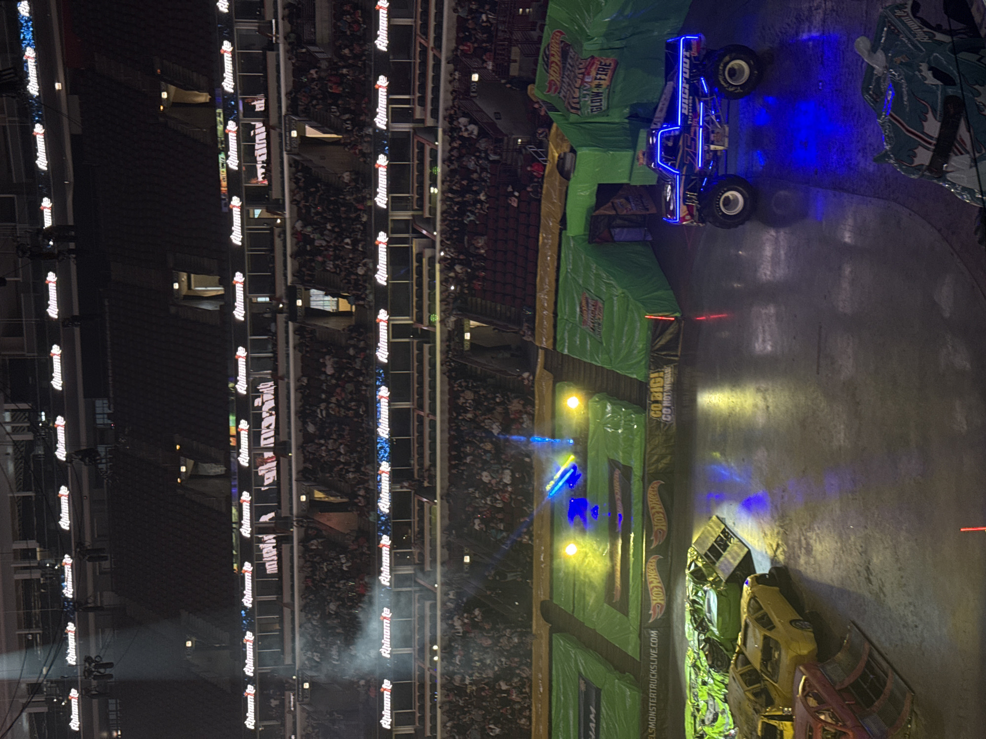 This is a high-angle, full shot of a monster truck arena in Chicago, United States of America, at night. The stadium is filled with a large audience seated in tiered bleachers that surround the arena floor. Above the crowd, numerous illuminated signs with white text on a black background, reading "Rhino-tie," are visible. The arena floor is covered in dirt, with several crushed cars arranged as obstacles. A prominent green-covered ramp structure is positioned on the left side of the arena. In the center-right of the arena, a blue monster truck with illuminated blue lights and "BIGFOOT" branding is parked. Another monster truck, partially visible in the foreground, has a blue and white patterned body. The overall atmosphere suggests a live event with dramatic lighting and a significant crowd presence.