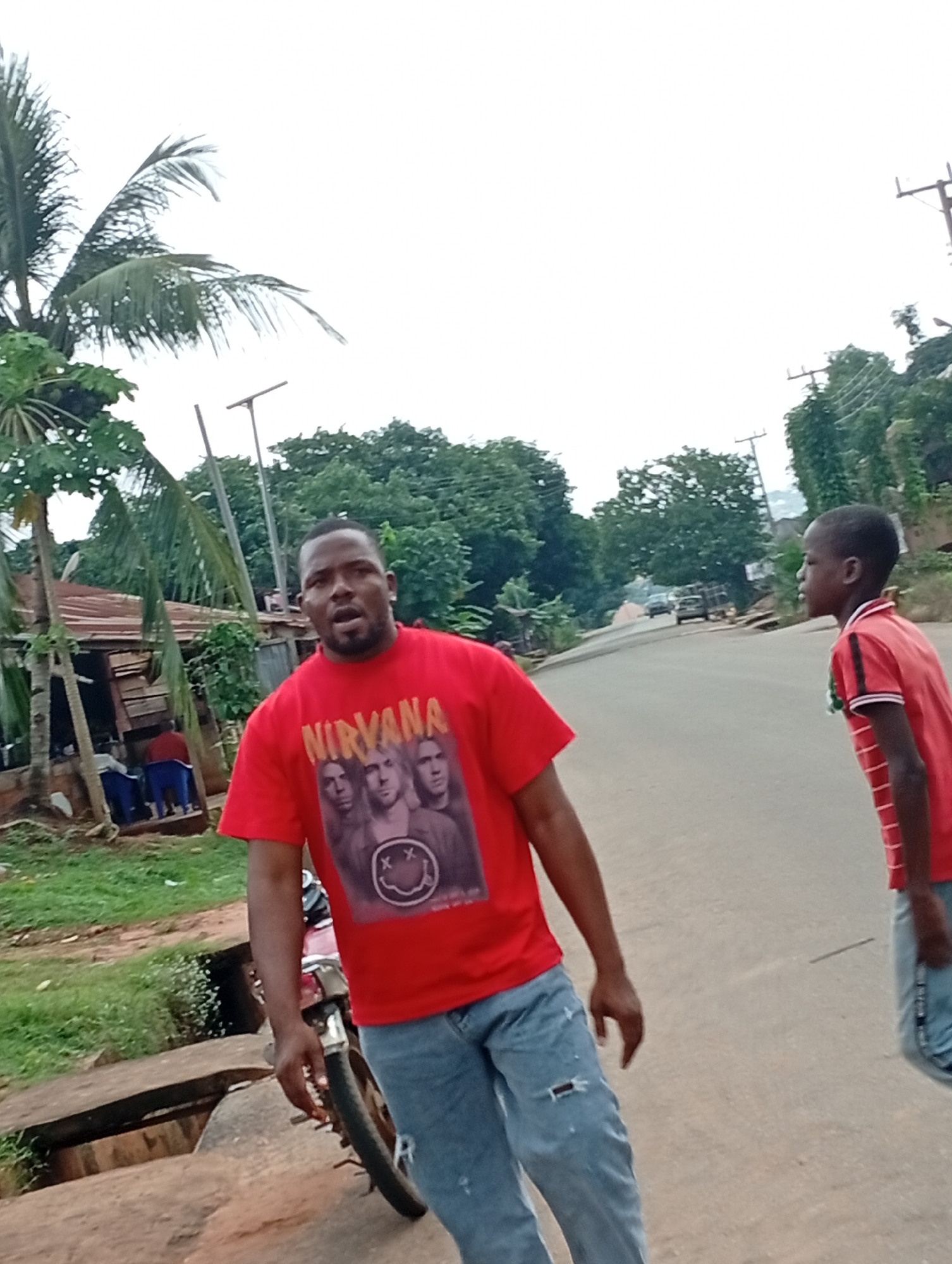 In Nsugbe, Nigeria, a man wearing a red t-shirt with a Nirvana band graphic and ripped blue jeans stands facing the camera. He is positioned in the foreground, slightly to the left of center. Behind him, to the right, a young boy in a red and white striped polo shirt walks away from the camera. The background features a road with a few vehicles, lush green trees, and some buildings with corrugated metal roofs. A motorcycle is partially visible in the lower left corner. The scene appears to be an outdoor, daytime setting.