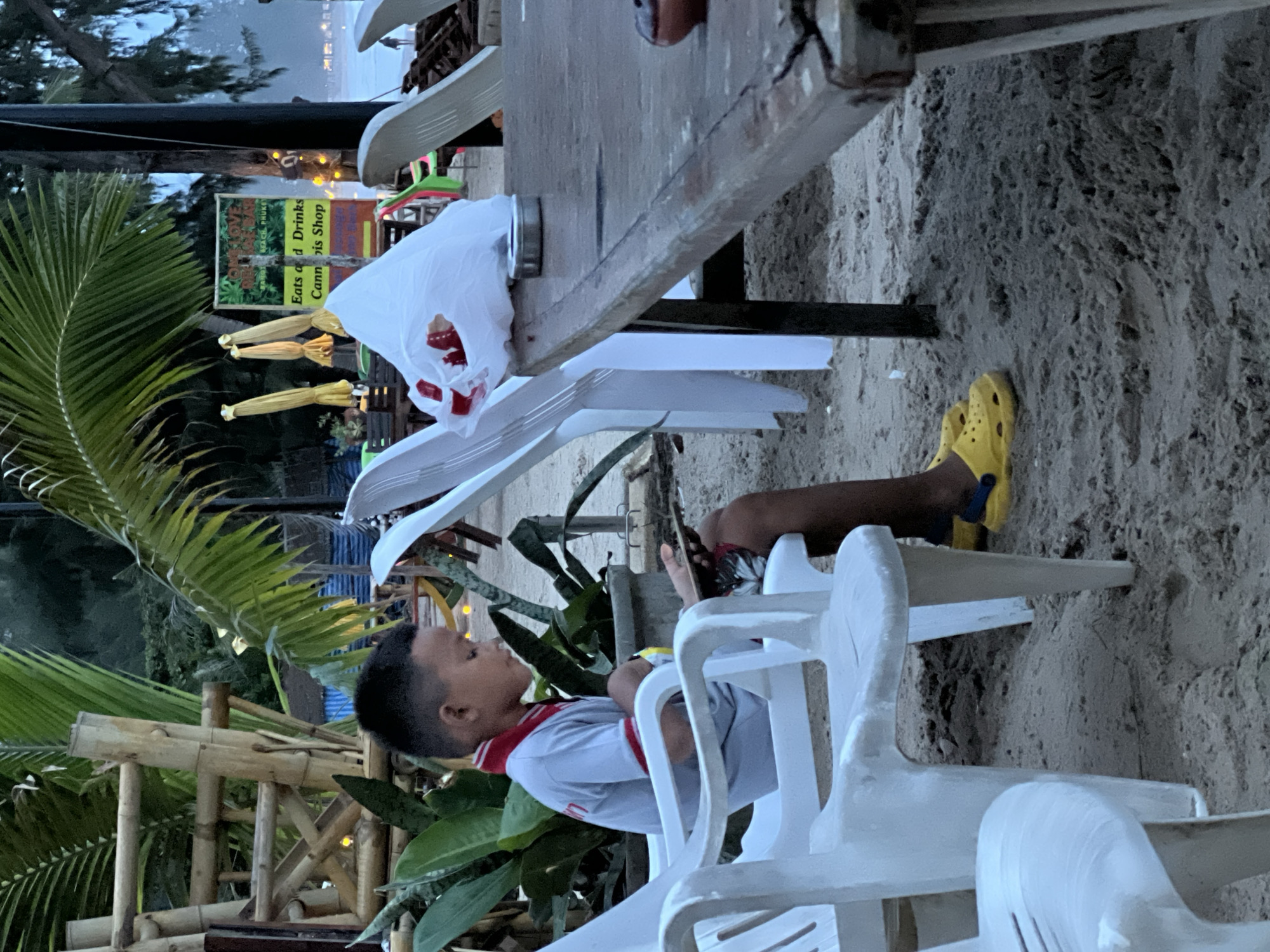 A slightly rotated medium shot captures a young boy sitting on a white plastic chair, engrossed in his cell phone. He sports a grey shirt with red trim and has a pair of yellow Crocs on his feet, which are resting on another white plastic chair. The setting appears to be outdoors on a sandy surface. In the background, a sign reading "Eats and Drinks Cannabis Shop" is visible, partially obscured by palm fronds and a string of lights. A wooden table and other chairs are also present. The location is an unspecified area in the city of Unknown, Unknown.