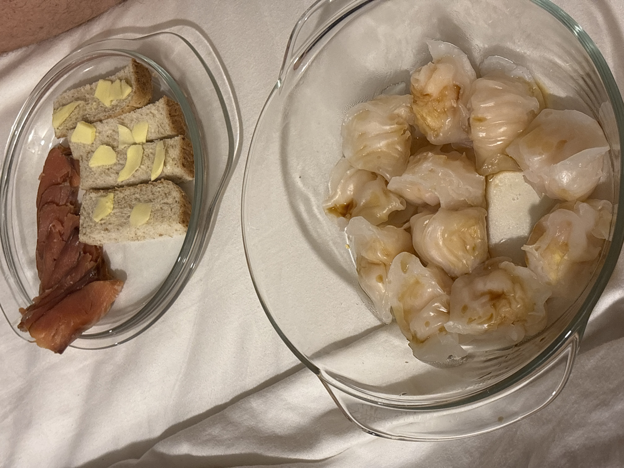 This image captures a close-up view of two clear glass bowls filled with food, set on a white, soft surface that appears to be bedding or a tablecloth. The scene takes place indoors, likely in a bedroom or hotel room in Choeng Thale, Thailand, creating a relaxed and intimate atmosphere.
The bowl on the left holds several slices of light-colored bread, possibly whole wheat or multigrain, each adorned with small, neatly placed pats of butter. Alongside the bread, a portion of thinly sliced, reddish-brown cured or smoked meat, resembling salmon or a similar deli meat, is arranged.
The bowl on the right contains numerous steamed dumplings, typical of Asian cuisine such as har gow (shrimp dumplings). These dumplings have a translucent, slightly pleated white skin, allowing glimpses of a lighter-colored filling within. Some dumplings show traces of a light brown sauce or liquid, indicating they might be served with a dipping sauce or have been steamed in a savory liquid.
A small section of a person's bare arm or leg is visible at the top left of the frame, suggesting an individual is present, possibly enjoying or about to consume the meal. The lighting is soft and ambient, indicative of an indoor setting, though it does not provide specific clues about the time of day. There is no visible text or explicit emotional display in the image.