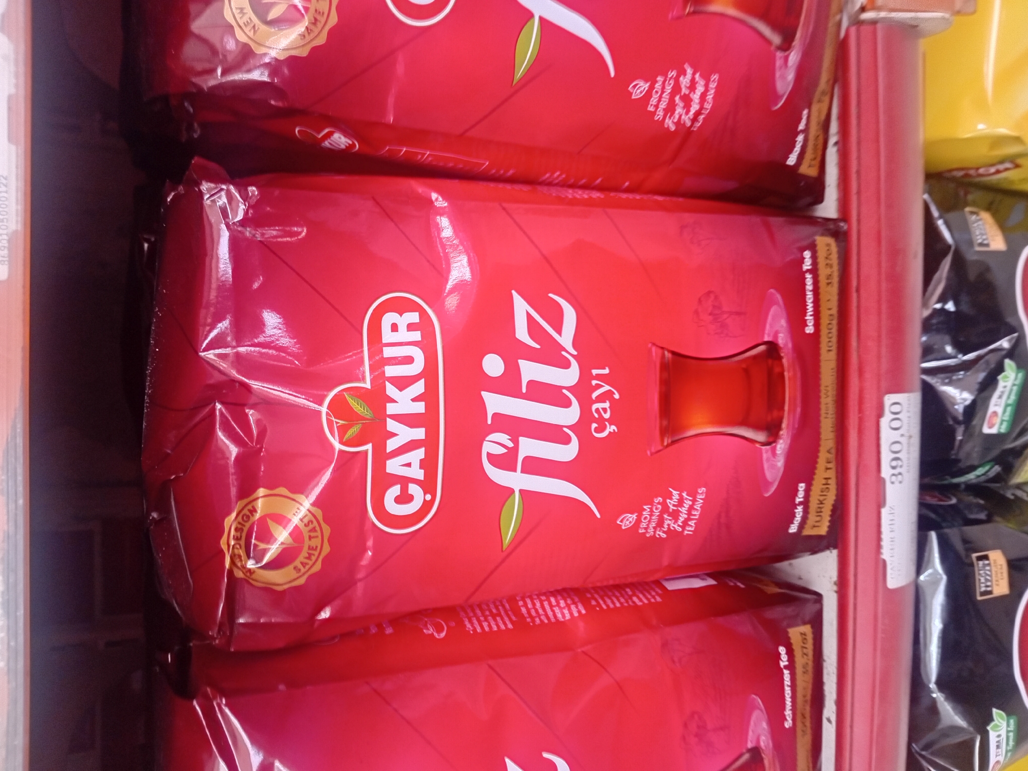A close-up, eye-level, indoor shot shows three large, red, plastic bags of Caykur Filiz brand tea on a red shelf in what appears to be a supermarket. The bag in the center is the most prominent, displaying the brand logo, the product name "filiz" in large white script, and "çay" in smaller white script below it. To the left and right of this central bag are similar, partially obscured bags of the same tea. The packaging features a circular seal with "NEW DESIGN" and "SAME TASTE" text around a leaf emblem. Below the main text on the central bag, there is a graphic of a traditional Turkish tea glass filled with tea, and text stating "FROM SPRING'S First And Freshest TEA LEAVES." The bottom of the bags show text like "Schwarzer Tee" and "Black Tea" with weight information. Below the shelf, a price tag reading "390,00" is visible. The lighting is even, and the focus is on the tea products. The scene is static, with no people visible. This scene is likely in a grocery store in Onikişubat, Turkey.