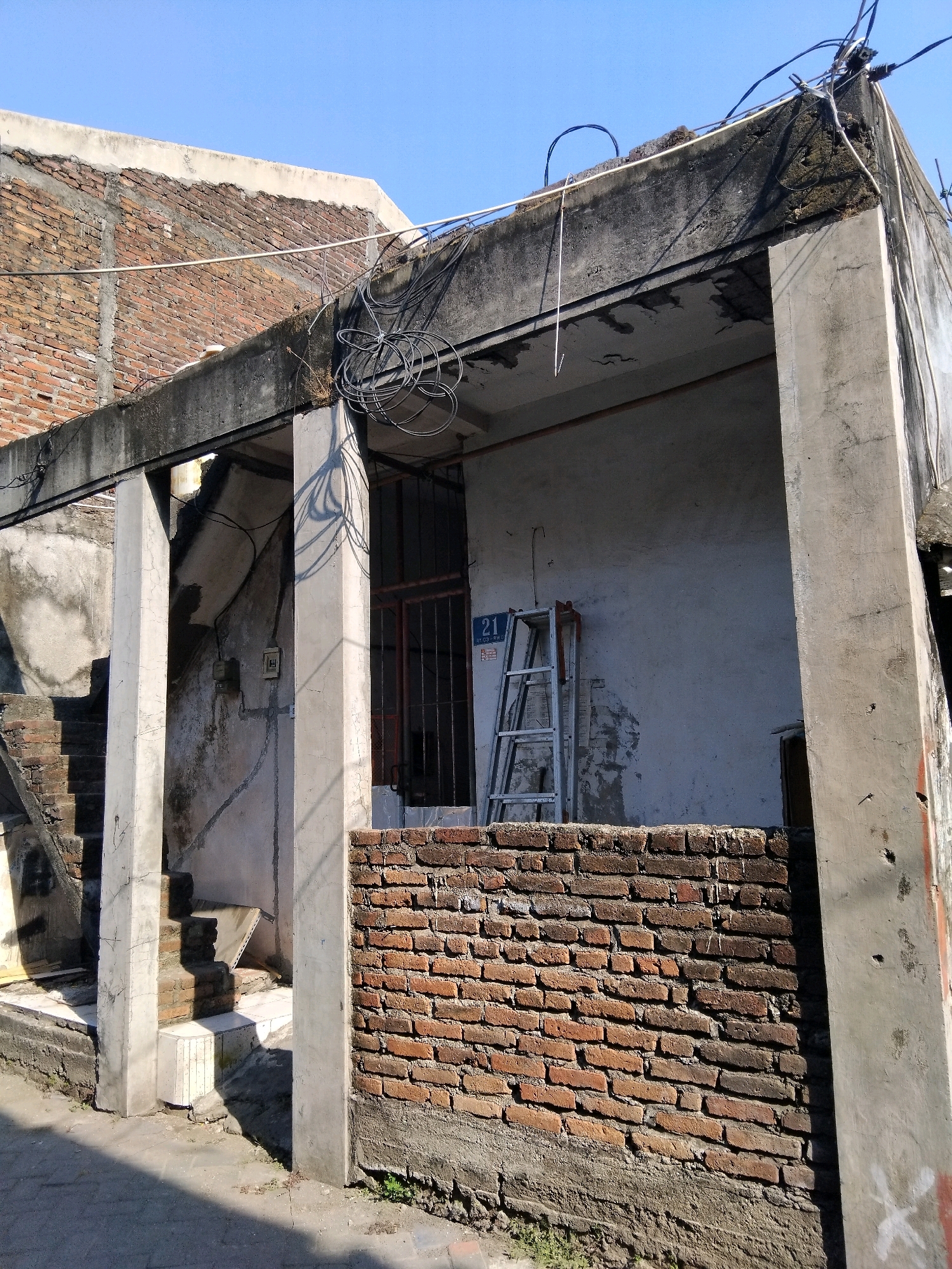 The image depicts a close-up of an old, somewhat dilapidated building with exposed brickwork and concrete elements, likely in Kepuh Kiriman, Indonesia. The foreground is dominated by a low brick wall. To the left, a set of concrete stairs is visible, partially obscured by shadows. The main structure features concrete columns supporting a roof overhang. The wall behind the columns is weathered and discolored, with visible electrical conduits and a junction box attached. A metal-framed window with bars is set into this wall. Leaning against the wall, to the right of the window, is a silver-colored ladder. Above the roofline, tangled electrical wires are prominent against a clear blue sky. The building appears to be in a state of disrepair, suggesting it is either under construction, renovation, or has been neglected. The scene is bathed in sunlight, casting distinct shadows. There are no people visible in the image.