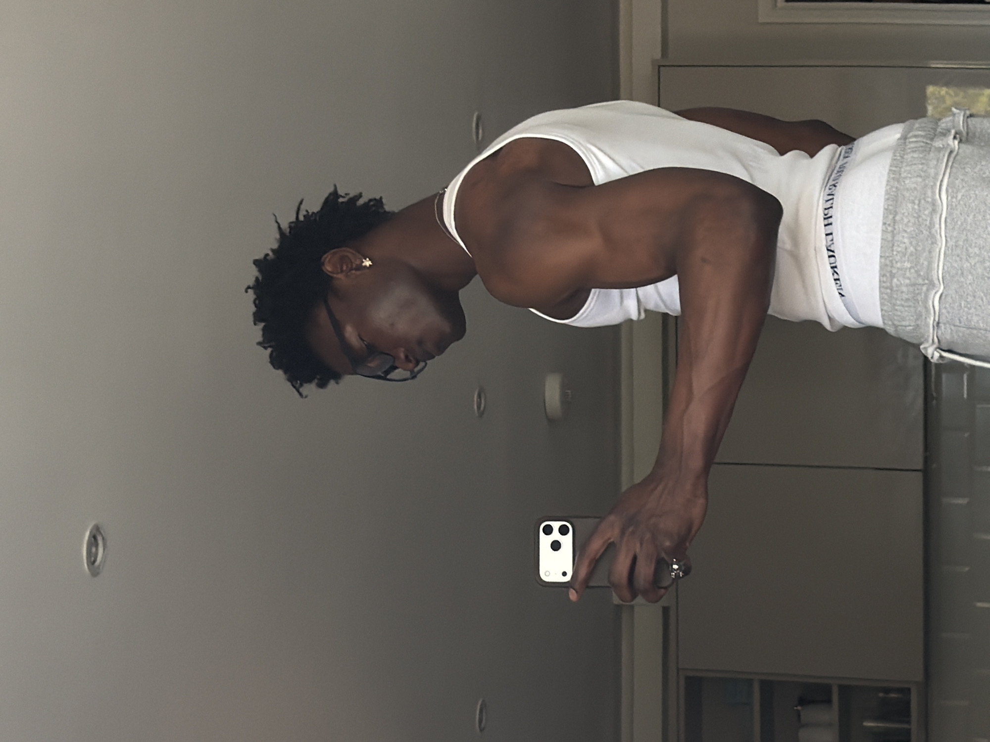 A person with dark skin, short dark curly hair, and glasses is depicted taking a mirror selfie indoors. They are wearing a white tank top and grey sweatpants, revealing a muscular physique in their arms and shoulders. A star-shaped earring is visible in their left ear, and a ring is on their right hand, which holds a smartphone (an iPhone with a triple camera setup). The person is looking down, focused on their reflection or the phone screen.
The background shows a light-colored wall with recessed lighting. On the right side, there are light grey cabinets or built-in furniture, suggesting an interior space like a bathroom or dressing room. The image is rotated 90 degrees clockwise from its natural upright orientation.
Notable details include the visible text "RALPH LAUREN" on the waistband of the person's underwear or sweatpants, which appears mirrored and upside down due to the mirror selfie. The indoor lighting is bright and even. The overall aesthetic of the room suggests a modern interior, consistent with an urban setting like London, United Kingdom. The person's expression is neutral.