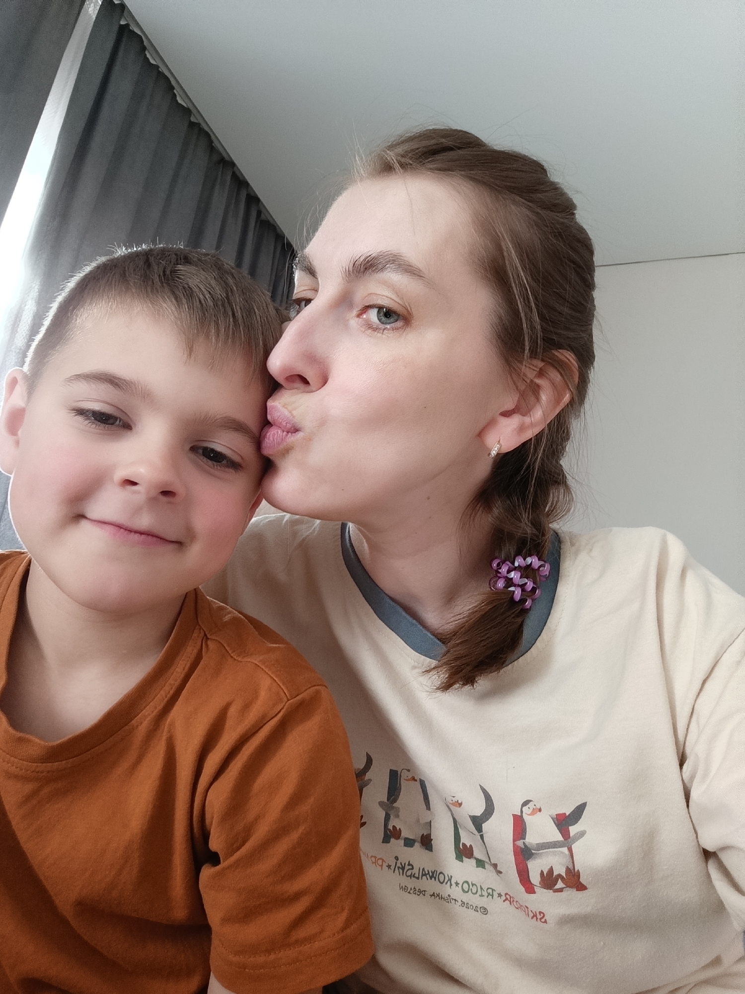 A selfie shot shows a woman kissing a young boy on the cheek. The boy has short brown hair and is wearing an orange t-shirt. The woman has brown hair pulled back in a braid, secured with a purple hair tie, and is wearing a light-colored t-shirt with penguin illustrations. The setting appears to be indoors, with a curtain partially visible in the background. The location is implied to be Chaikovsky, Russia.