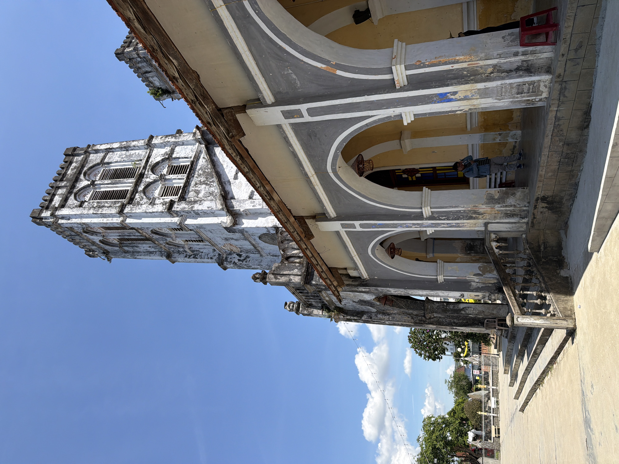A wide-angle, eye-level outdoor shot captures the exterior of a weathered church in Hội Tín, Vietnam. The church features a tall, stone bell tower with arched windows and a crenellated top, showing signs of age and neglect with peeling paint and cracks. To the right of the tower, a covered walkway with arched openings leads into the building. The walkway is supported by columns and features a yellow-painted interior. A few people are visible: one man in a denim jacket stands inside the arched walkway, appearing to be on his phone, while another person in darker clothing stands further inside. A red plastic stool is placed outside the walkway. In the foreground, a wide, concrete staircase with a stone balustrade leads up to the church entrance. The ground in front of the church is paved concrete, with some bare ground and greenery visible further in the distance. A string of small lights hangs across the scene under a bright blue sky with scattered white clouds. The overall impression is of an old, historical religious structure in a tropical climate.