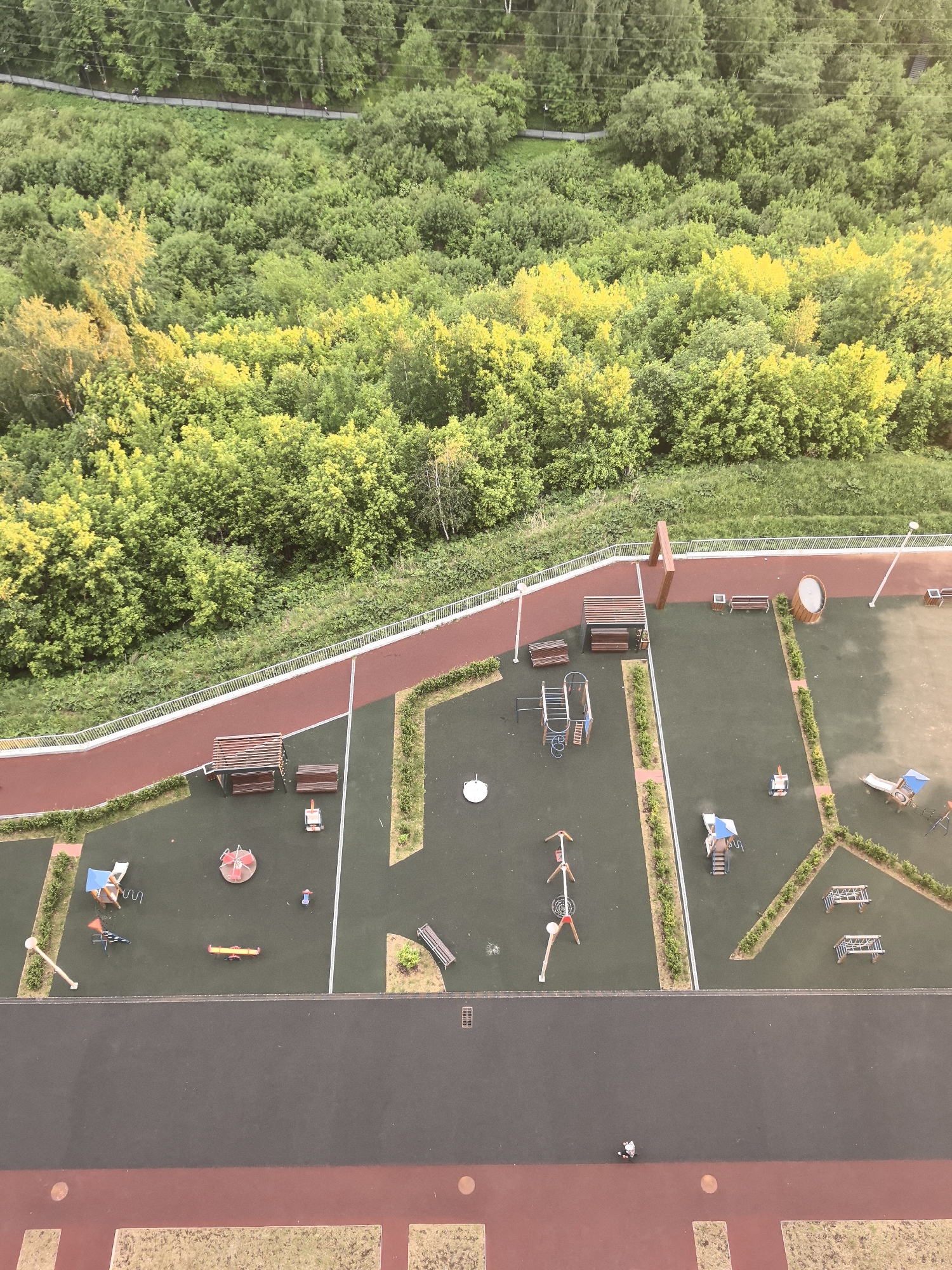 The image depicts an elevated view of a playground area in Perm, Russia, adjacent to a forested area. The playground contains various pieces of equipment including swings, slides, benches, and climbing structures. The play area is separated into distinct sections delineated by hedges and pathways. A dark grey paved area is visible at the bottom of the image, potentially a roadway or walkway bordering the playground. A red pathway runs along the edge of the playground, bordering the forest. The forest is dense with primarily green foliage. A footbridge is faintly visible in the background above the forest, indicating human activity in the surrounding natural area. The lighting indicates the photograph was taken during daylight. A person is visible on the ground.
