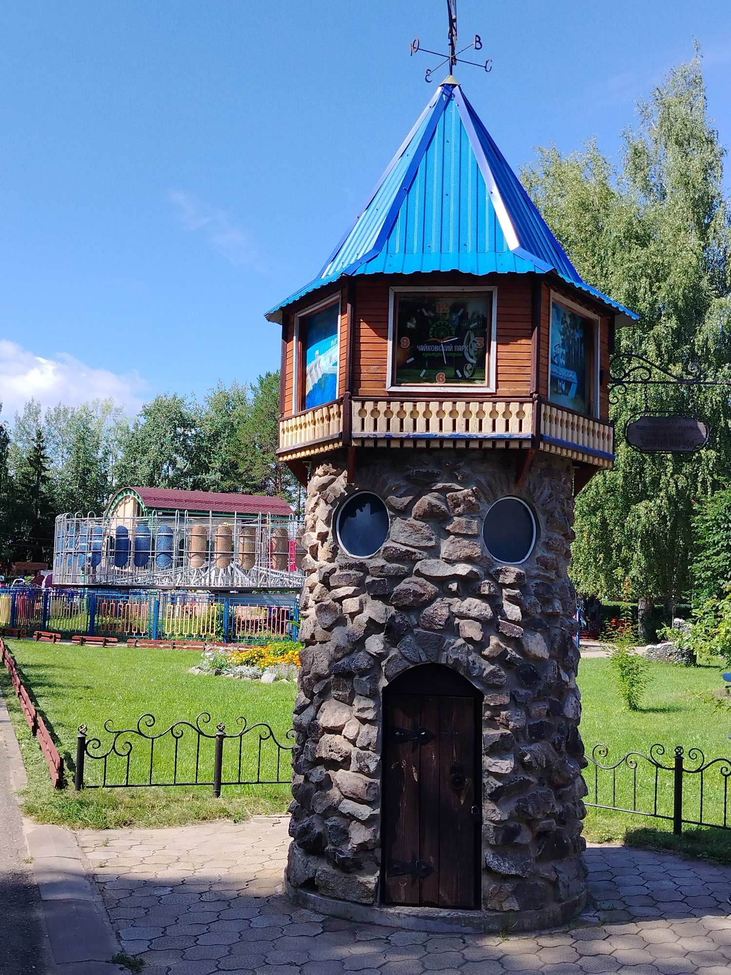 This is a photograph of a whimsical, stone tower with a blue, peaked roof, located in a park. The tower has a wooden door at its base and two circular windows. The upper section of the tower is made of wood and features a balcony with a decorative railing. There are windows on the upper section that appear to be decorative displays, one of which shows a clock face with Russian text that translates to "Chaikovsky Park". A weather vane is visible on the very top of the tower.
The tower is situated in a park with a grassy area, flower beds, and a paved walkway. In the background, there is a colorful playground structure with a Ferris wheel and a red-roofed building. Trees and a bright blue sky with some clouds form the backdrop. The scene suggests a sunny day in a public recreational space. The presence of the "Chaikovsky Park" text on the tower strongly indicates the location.